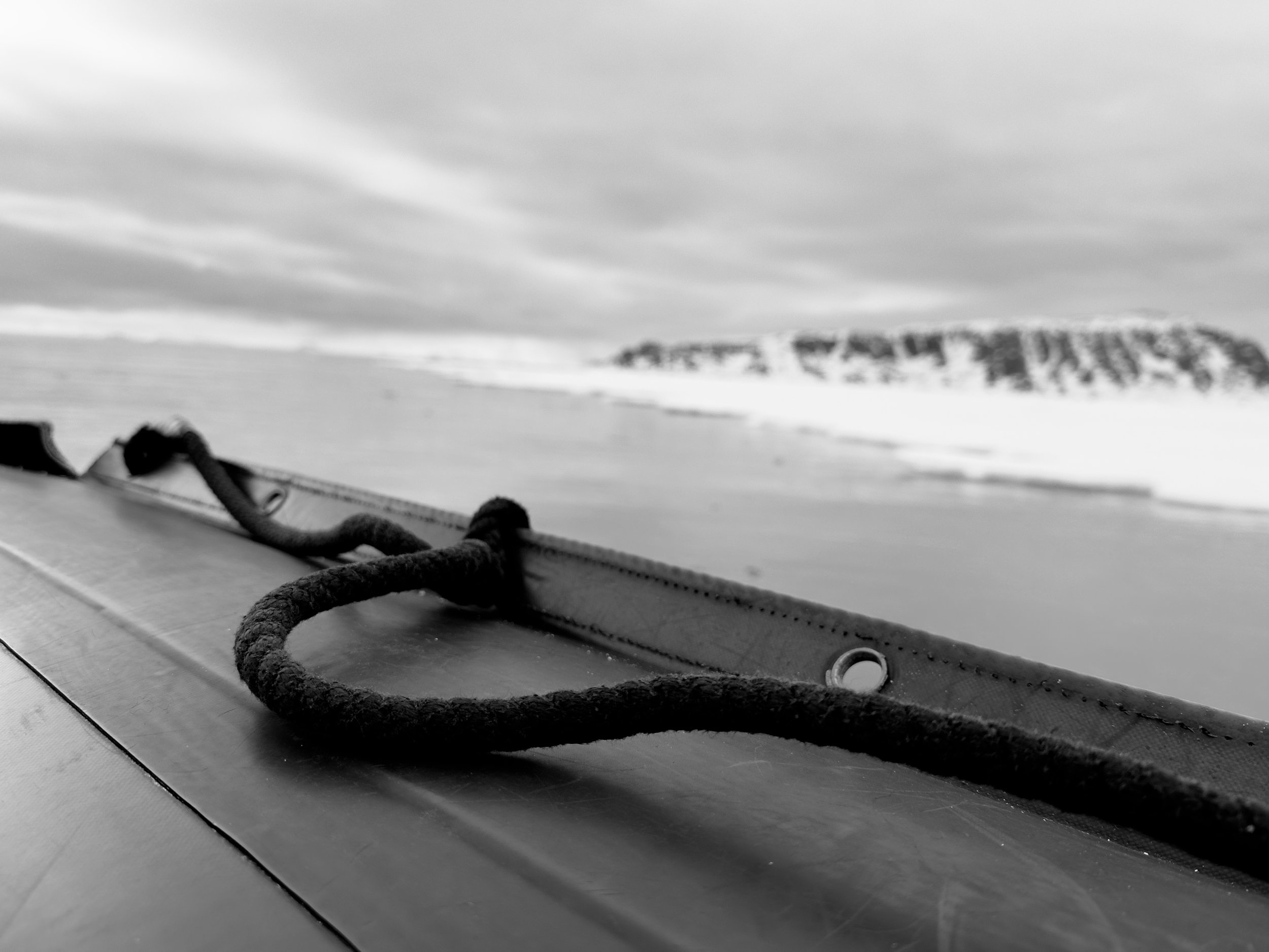 Close-up of a boat's deck with a rope tied to a cleat, overlooking a snowy landscape with distant hills and cloudy sky.
