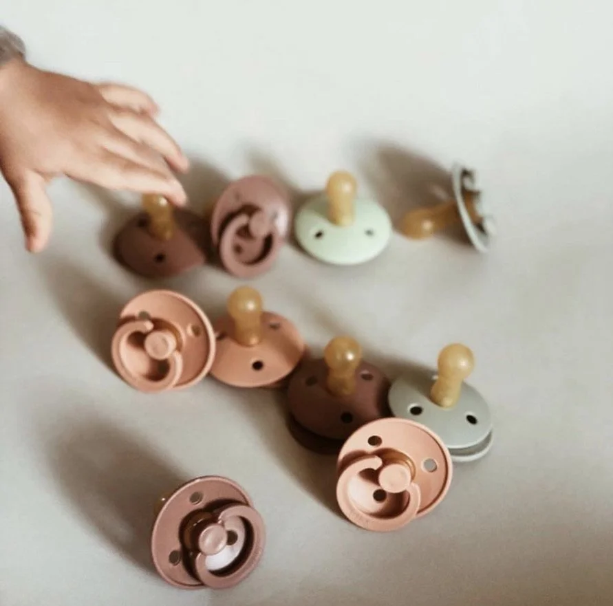 Hand reaching out toward colorful pacifier covers with plastic handles on a white surface.