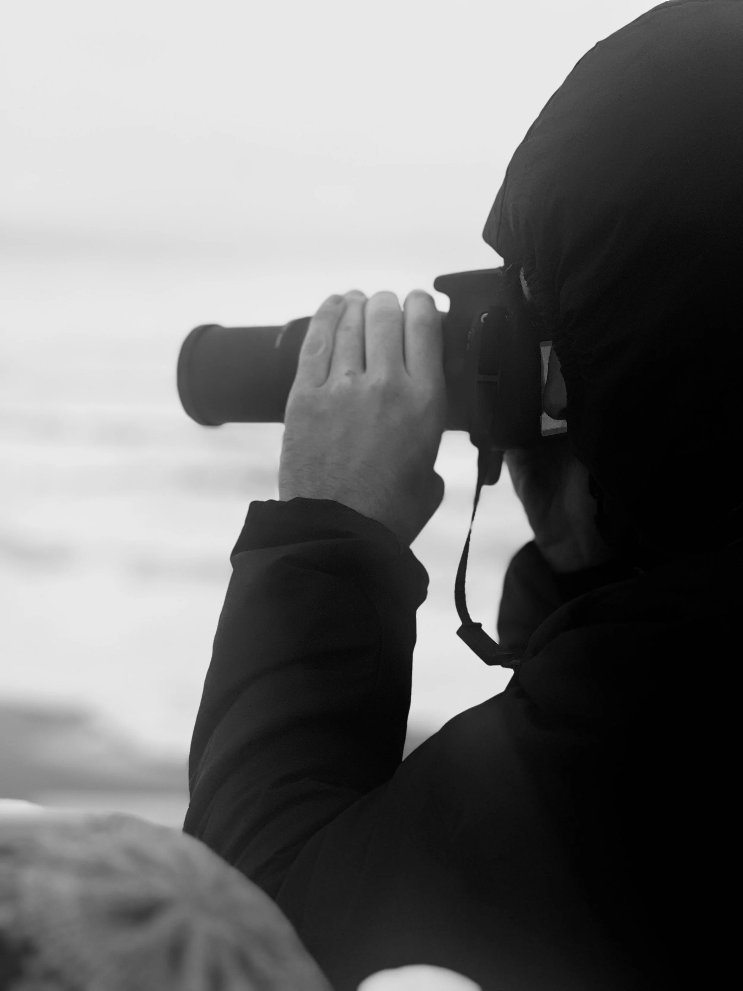 Person in a hooded jacket taking a photograph with a camera outdoors in black and white.