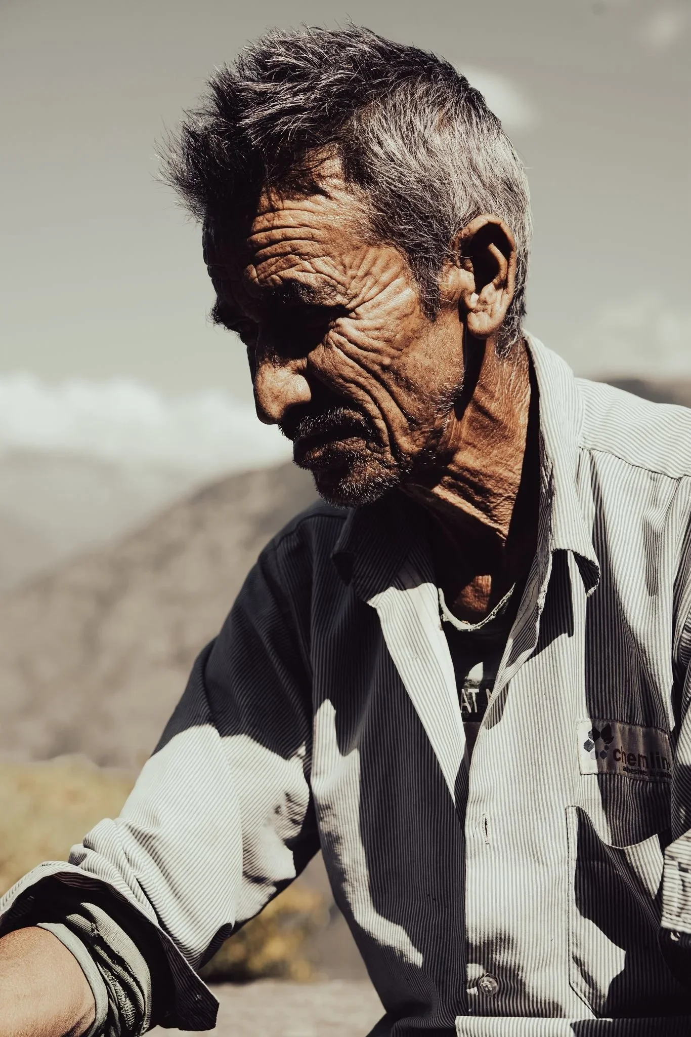 A close-up side profile of an elderly man with gray hair, deep wrinkles, and a white beard wearing a striped shirt, outdoors with blurry background.