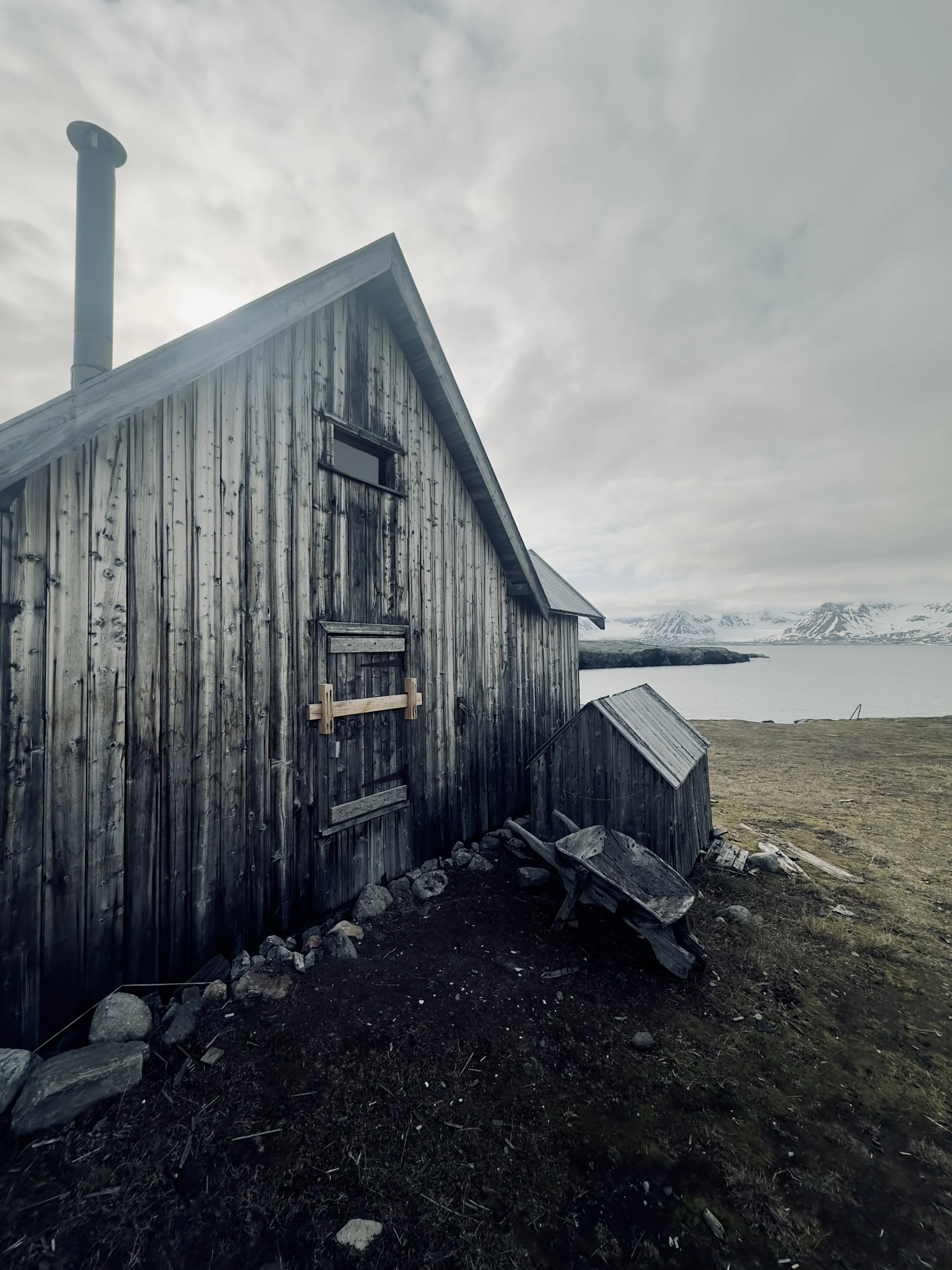 A weathered wooden building near a body of water with snow-capped mountains in the distance.