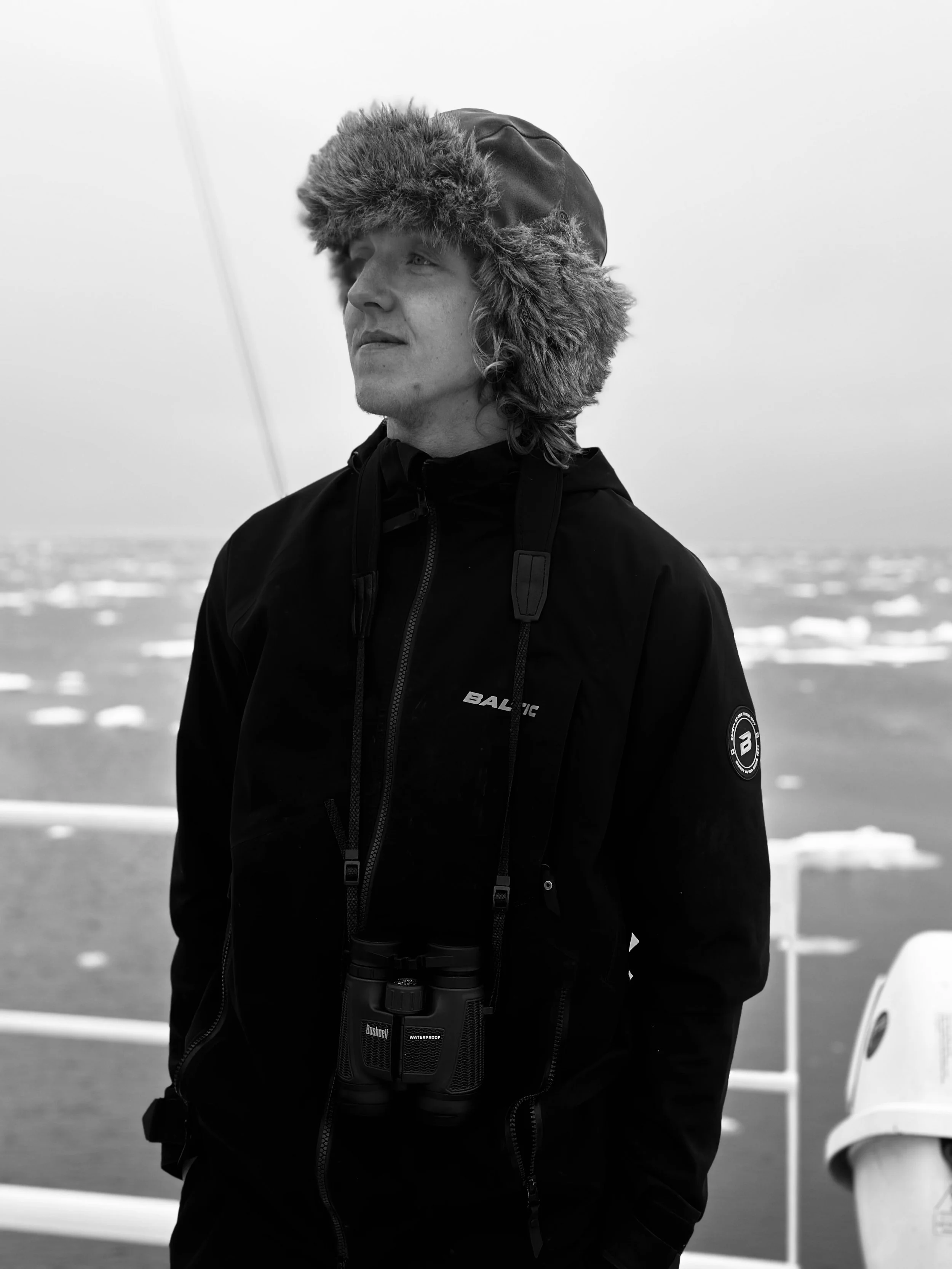A person wearing a warm hat with fur trim and a dark jacket with binoculars hanging around their neck, standing outdoors in a cold, icy landscape.