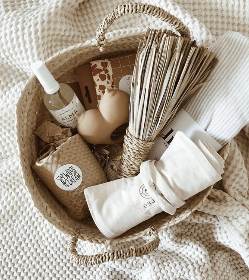 A woven basket filled with various beige and neutral-colored household items, including a spray bottle, candles, tissue paper, cloth napkins, and a wrapped gift, set on a textured beige blanket.