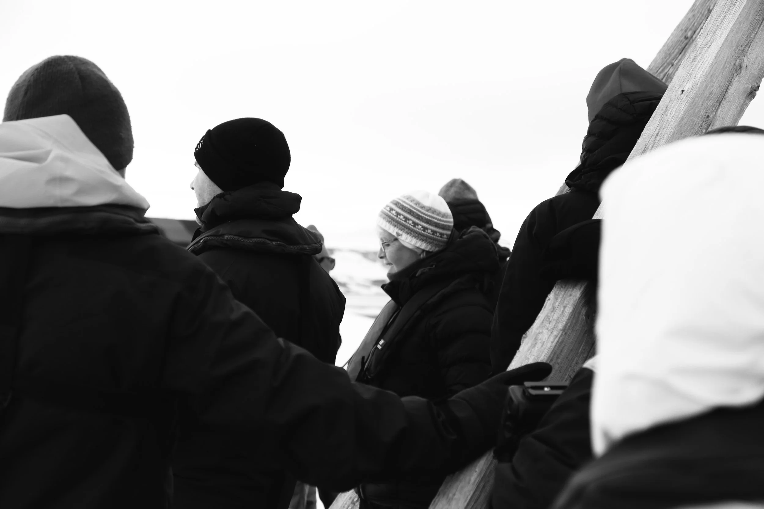Group of people dressed warmly, standing outdoors on a cold day. One woman with glasses and a patterned knit hat is primarily visible between other individuals wearing dark jackets and knit hats. They are near a large wooden structure.