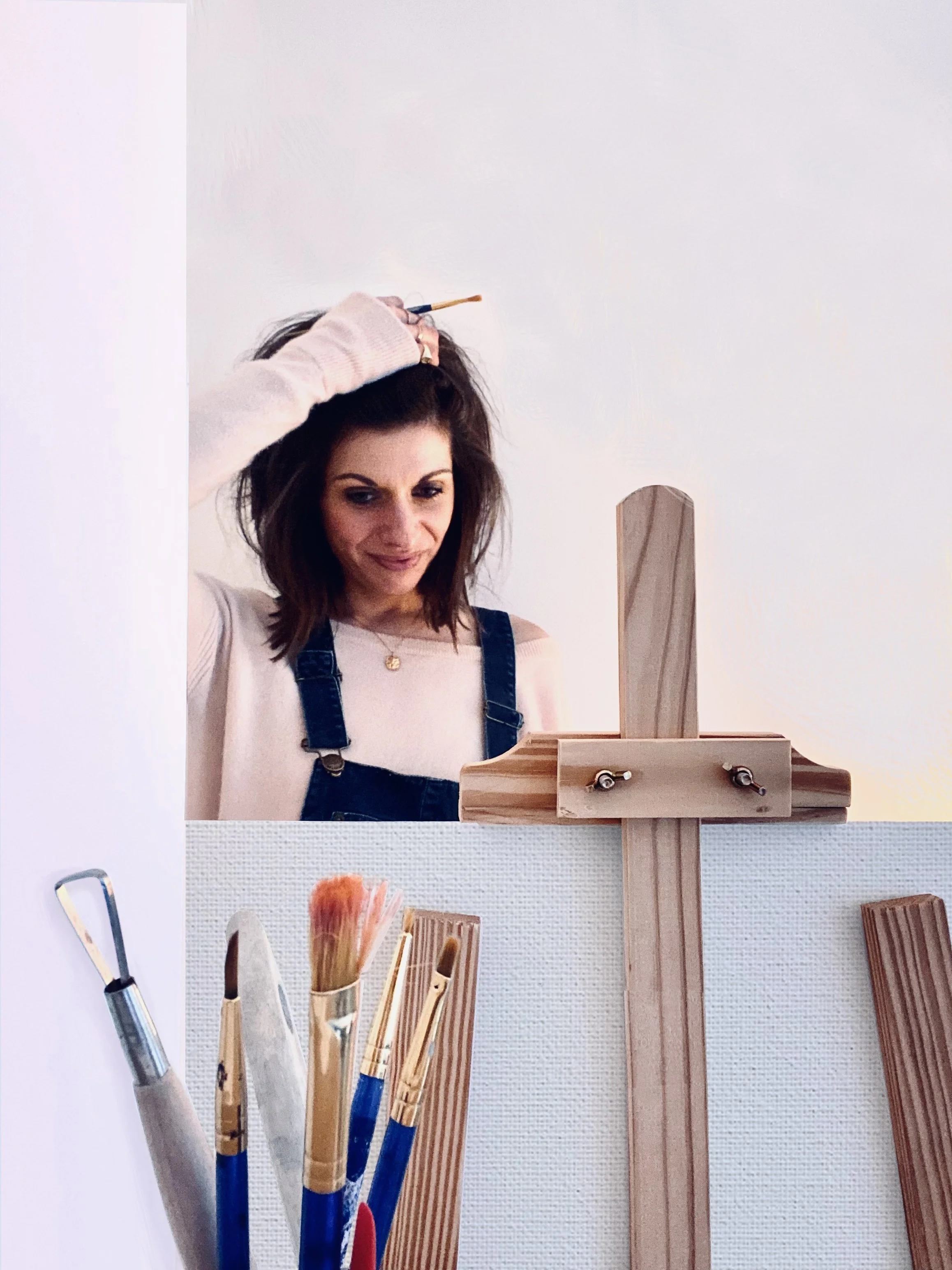 A woman with shoulder-length brown hair and a beige sweater looking down at a canvas while standing behind an easel. She is standing in front of a plain white wall, with art brushes and tools on a table in front of her.