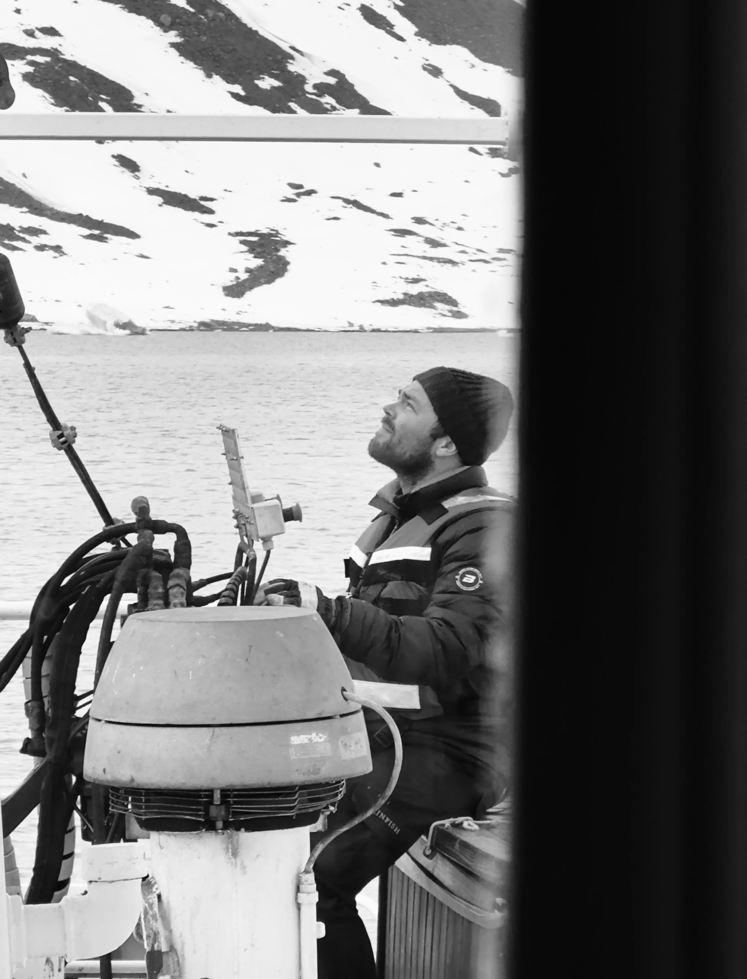 A man wearing a beanie and a jacket on a boat, looking up, with snowy mountains in the background.