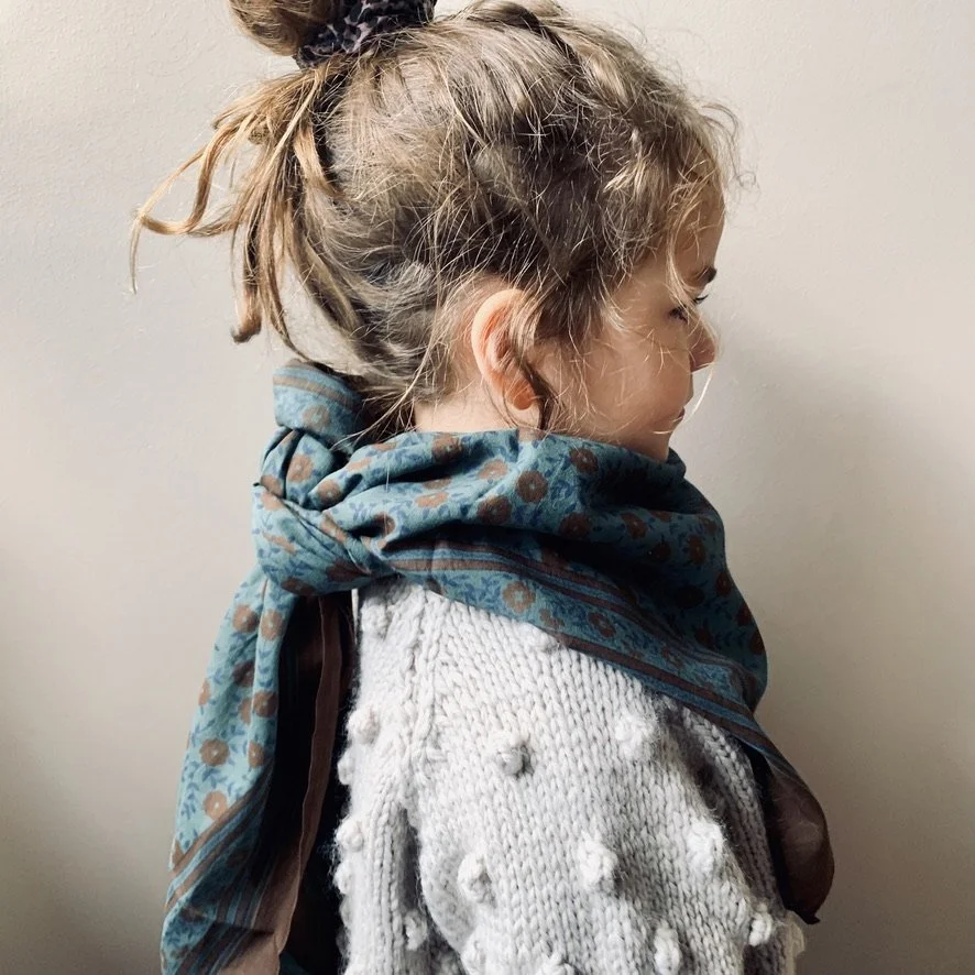 Side profile of a young girl with light brown hair tied up in a ponytail, wearing a white knitted sweater with pom-poms, and a blue patterned scarf.