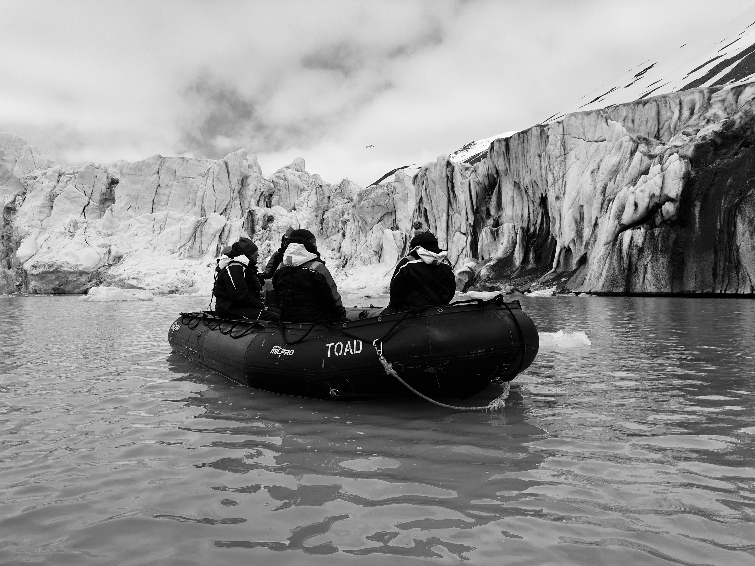 Four people in winter gear sitting in an inflatable boat on cold water, with glaciers and icy cliffs in the background.