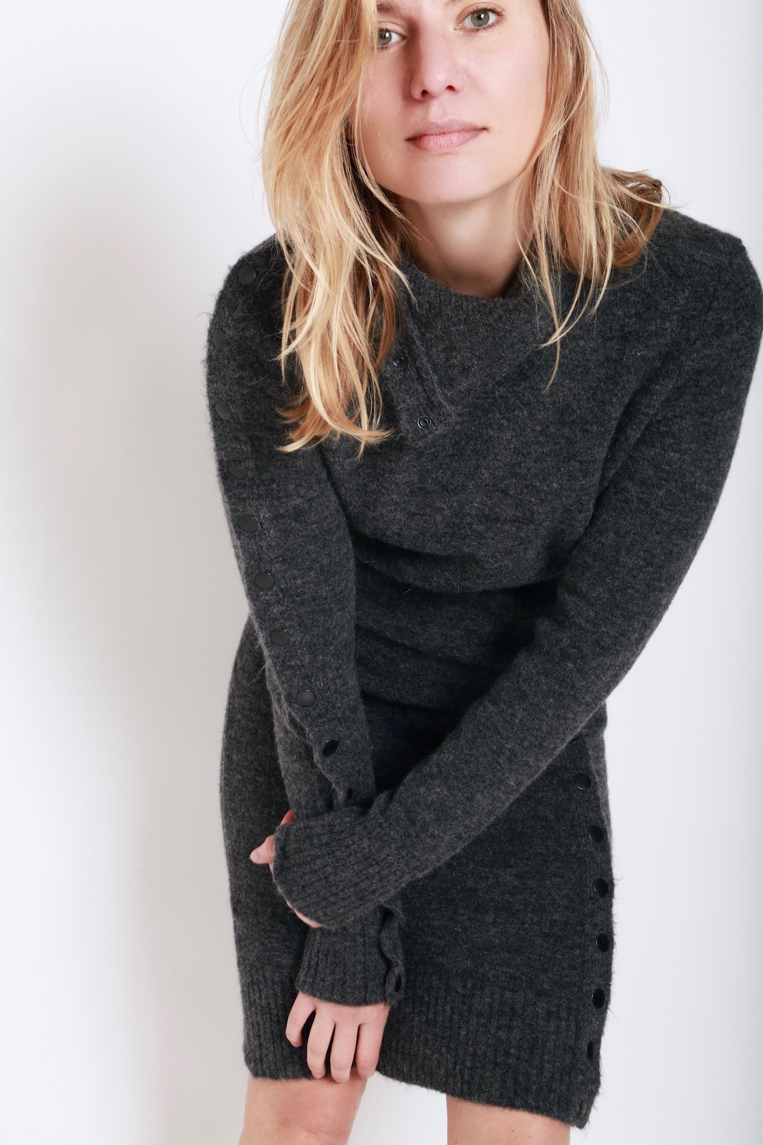 A woman with blonde hair wearing a dark gray sweater and matching skirt standing against a white background.
