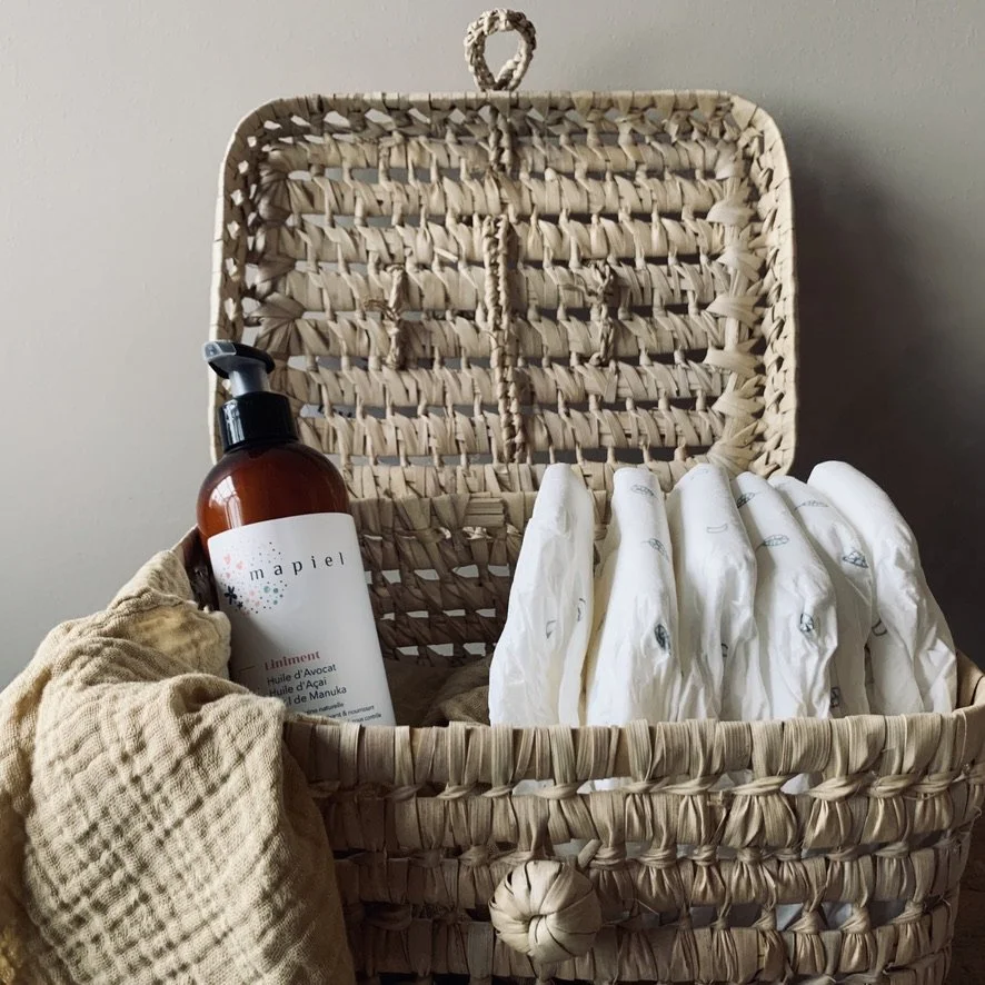A woven basket containing a brown bottle of hair or body oil, a beige cloth, and several white sanitary pads or cloths. The basket is open with a handle on top, placed against a plain light-colored wall.