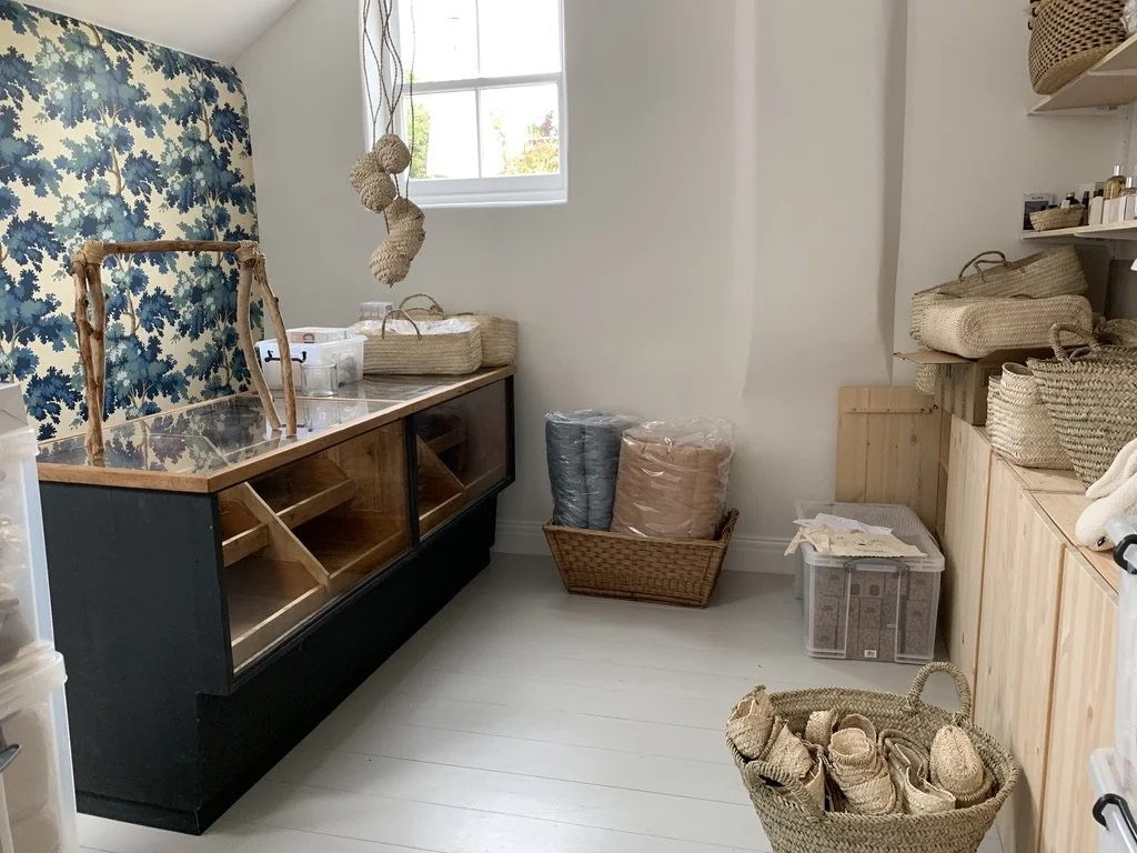 A small room with a window, covered with white walls and floor, containing shelves with woven baskets, a glass display case, and rolls of fabric or rugs.