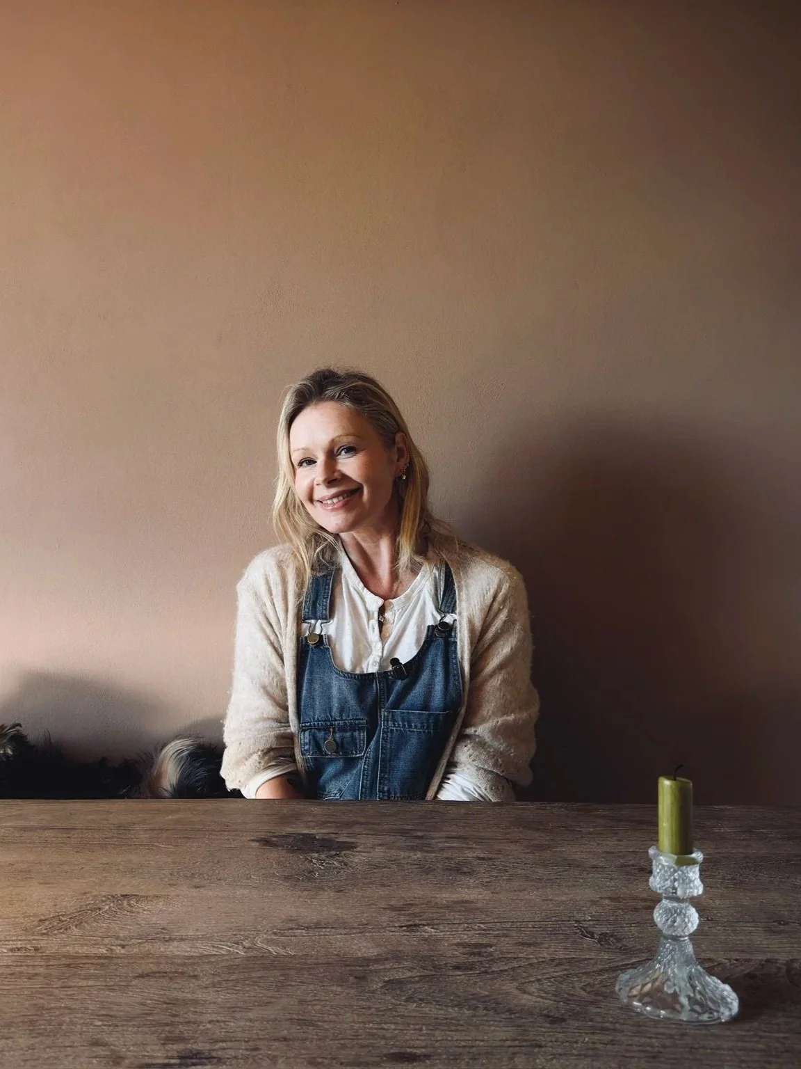 A woman with blonde hair smiling and sitting at a wooden table, wearing a cream sweater and denim overalls, with a green candle in a glass holder on the table.