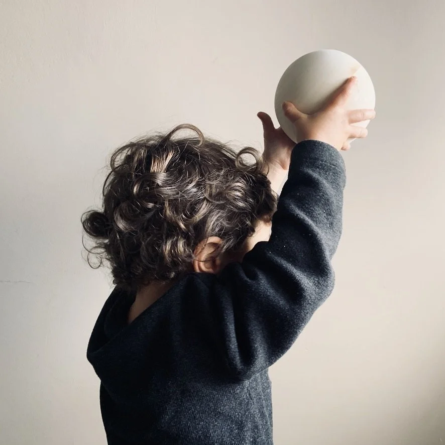 A child with curly hair holding up a round white object against a plain background.