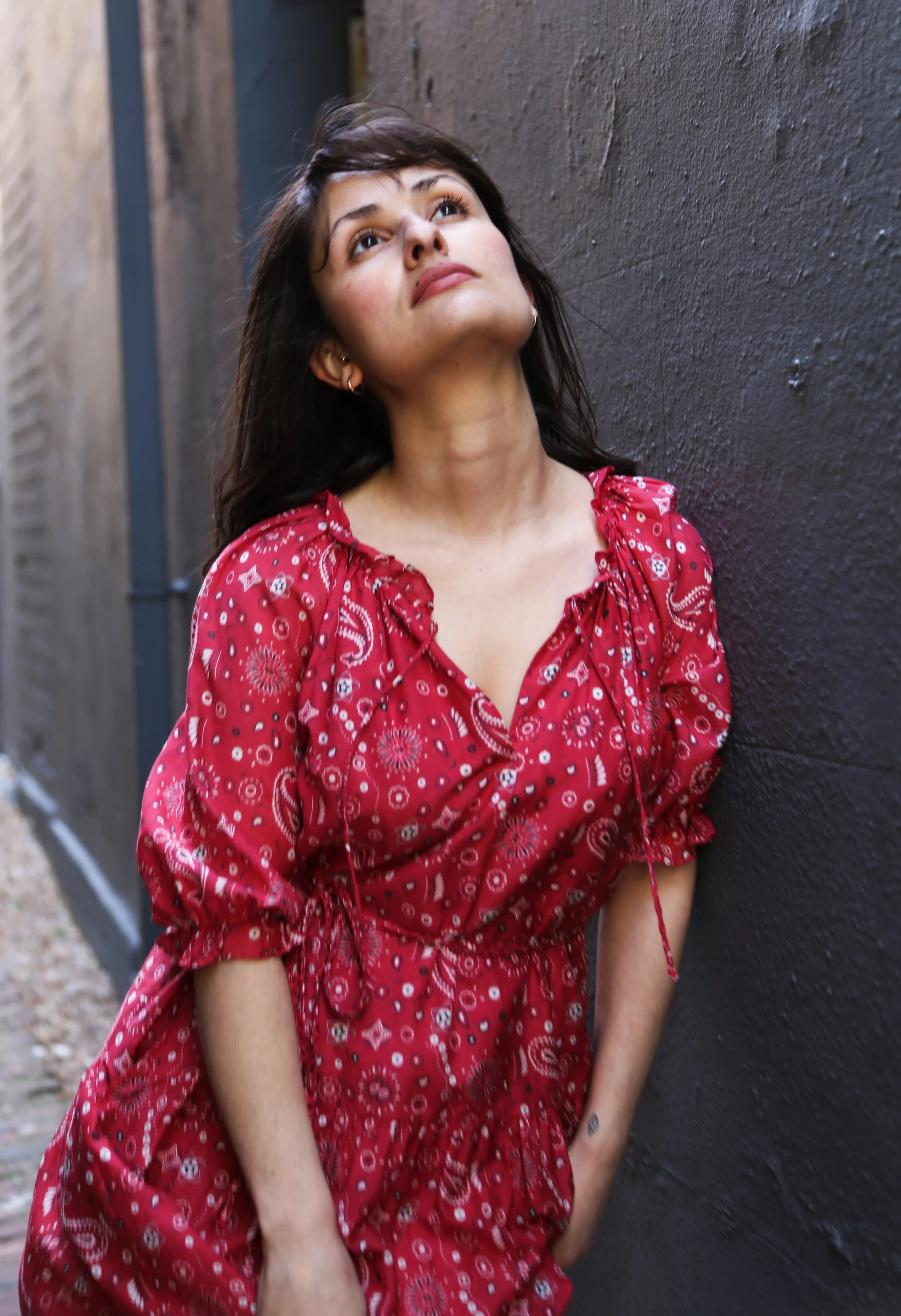 A woman in a red patterned dress leaning against a dark wall and looking upwards.