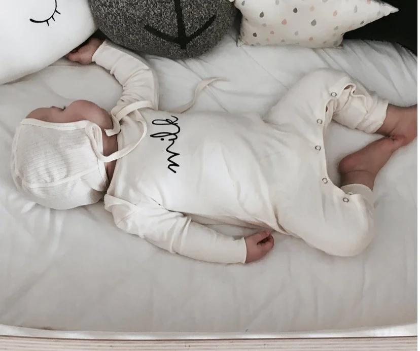 A sleeping baby dressed in white pajamas and a matching hat, lying on a bed surrounded by plush pillows.