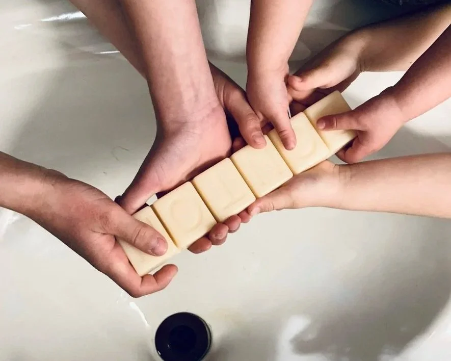 Multiple hands holding a row of soap bars speller spelling 'LOVE' over a sink.