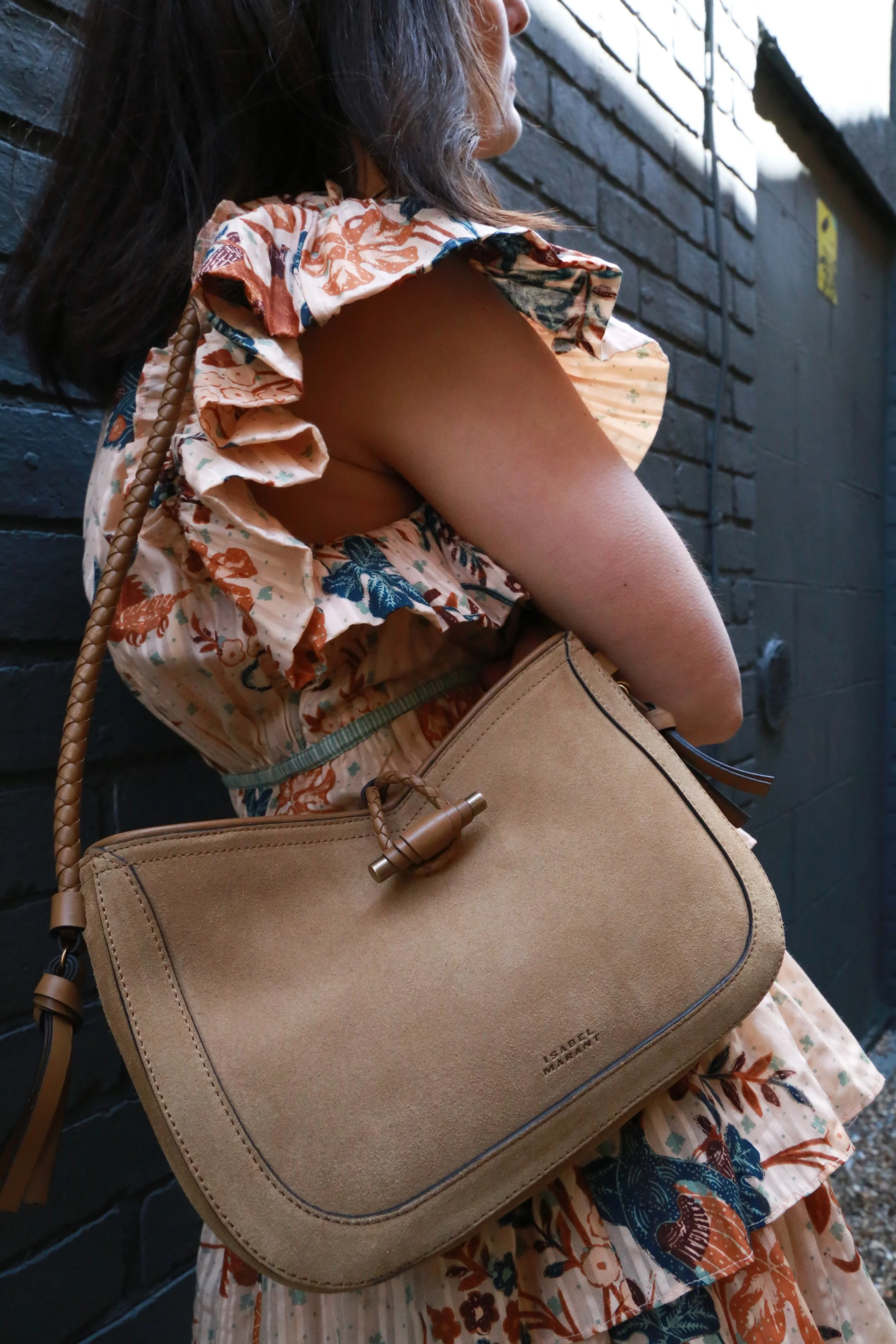 A woman standing against a dark brick wall, wearing a floral dress and carrying a tan shoulder bag.