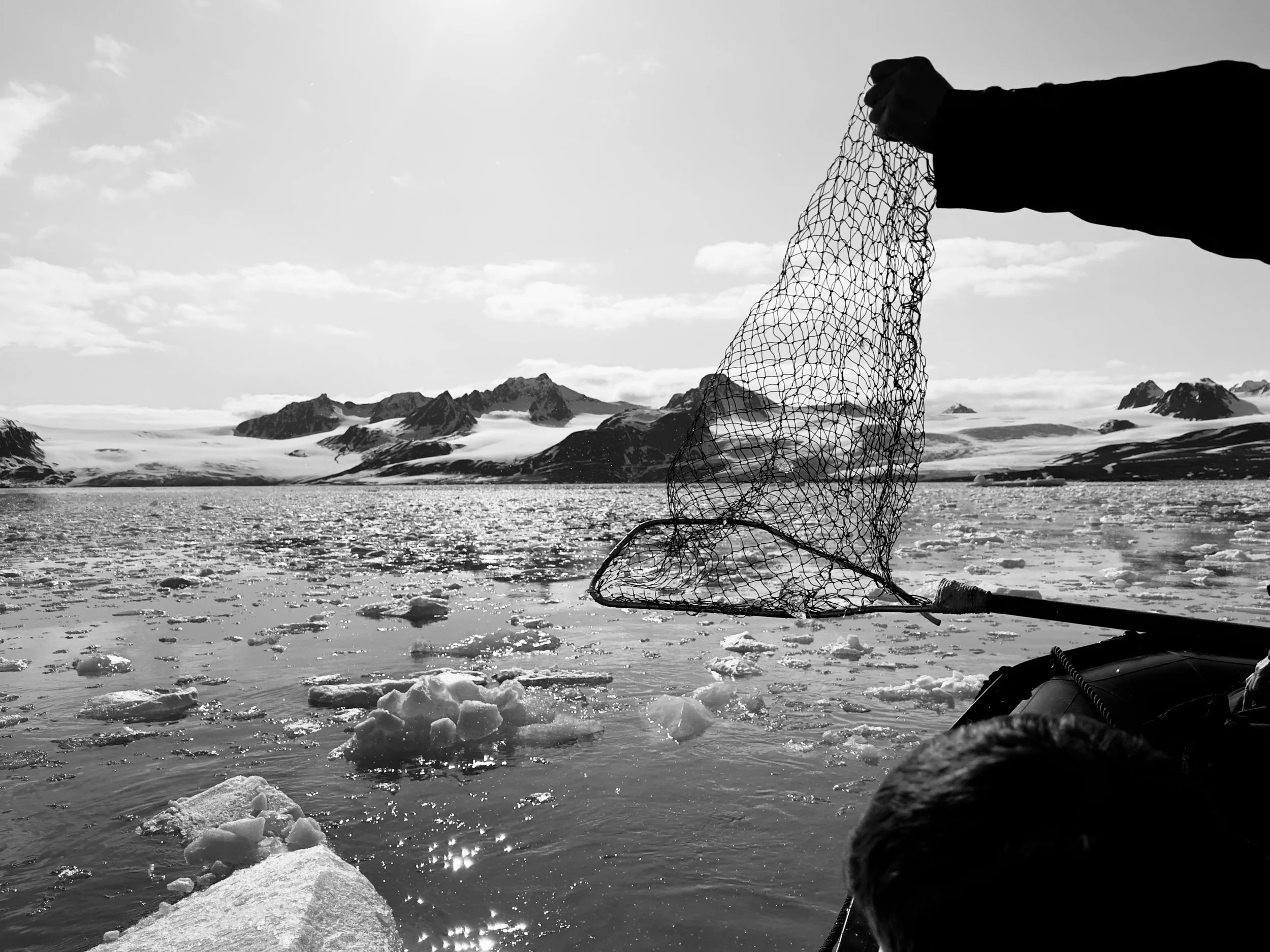 A person holding a fishing net over icy water in a mountainous, snowy landscape in black and white.