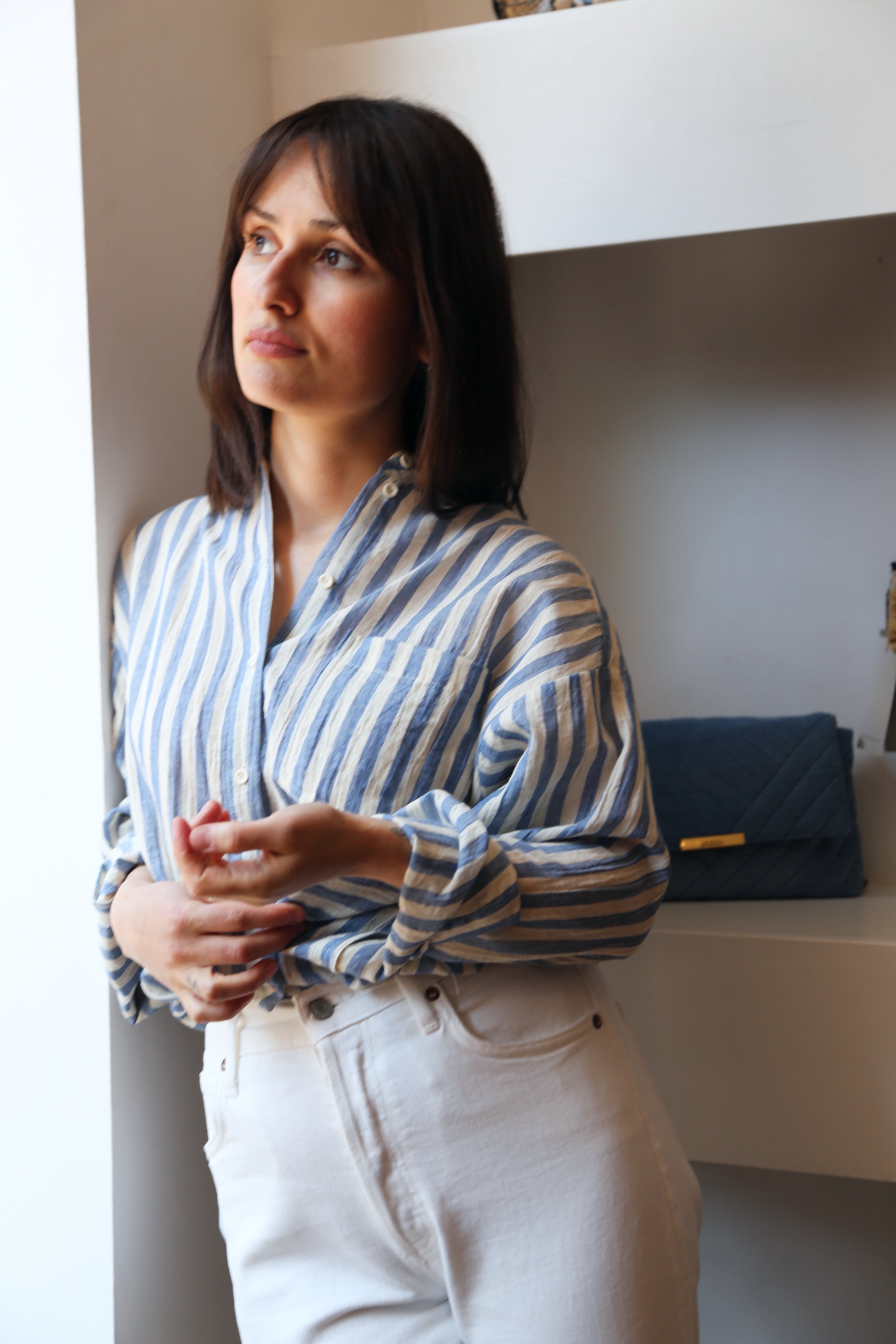 A woman with shoulder-length dark hair wearing a striped blue and white shirt, standing near a window with natural light, with a dark blue quilted clutch on display behind her.