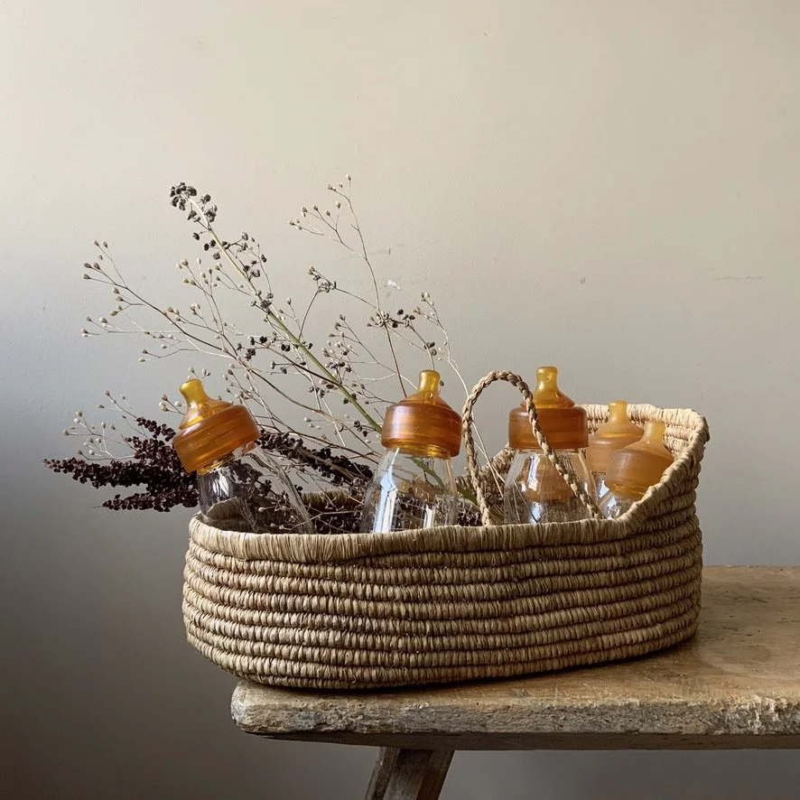 A woven basket containing dried plants and empty baby bottles with orange caps, placed on a wooden surface.