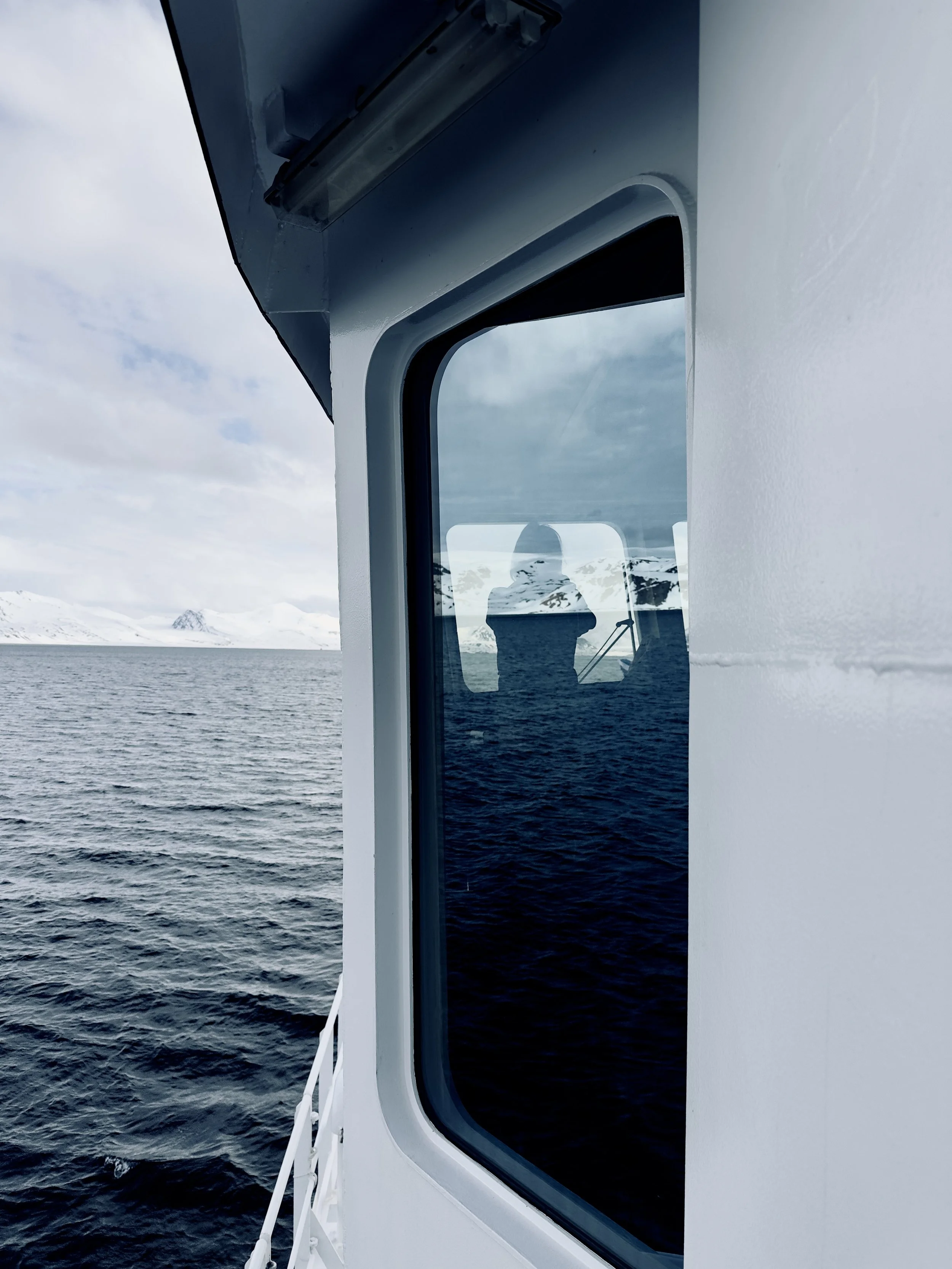 View of the ocean through a ship window, with a reflection of a person taking the photo, and snow-covered mountains in the background.