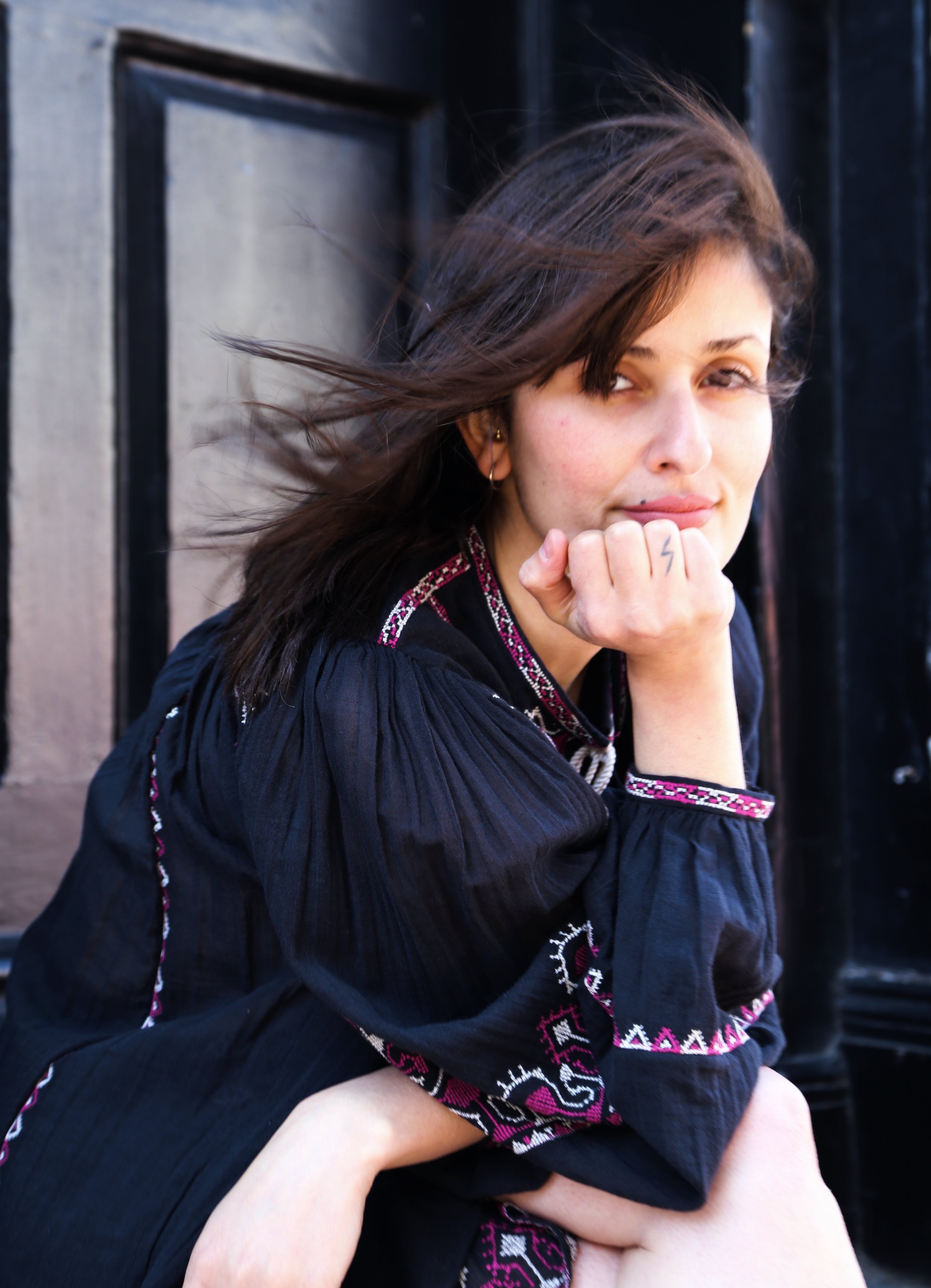 A woman with brown hair, wearing a black embroidered dress, resting her chin on her hand, looking at the camera, sitting outdoors near a black door.