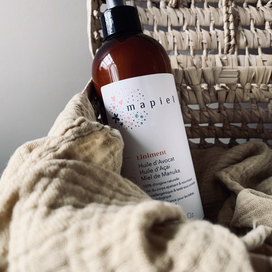 A brown bottle of Mapiel brand natural skincare product with a white label, resting on beige fabric with a woven basket in the background.