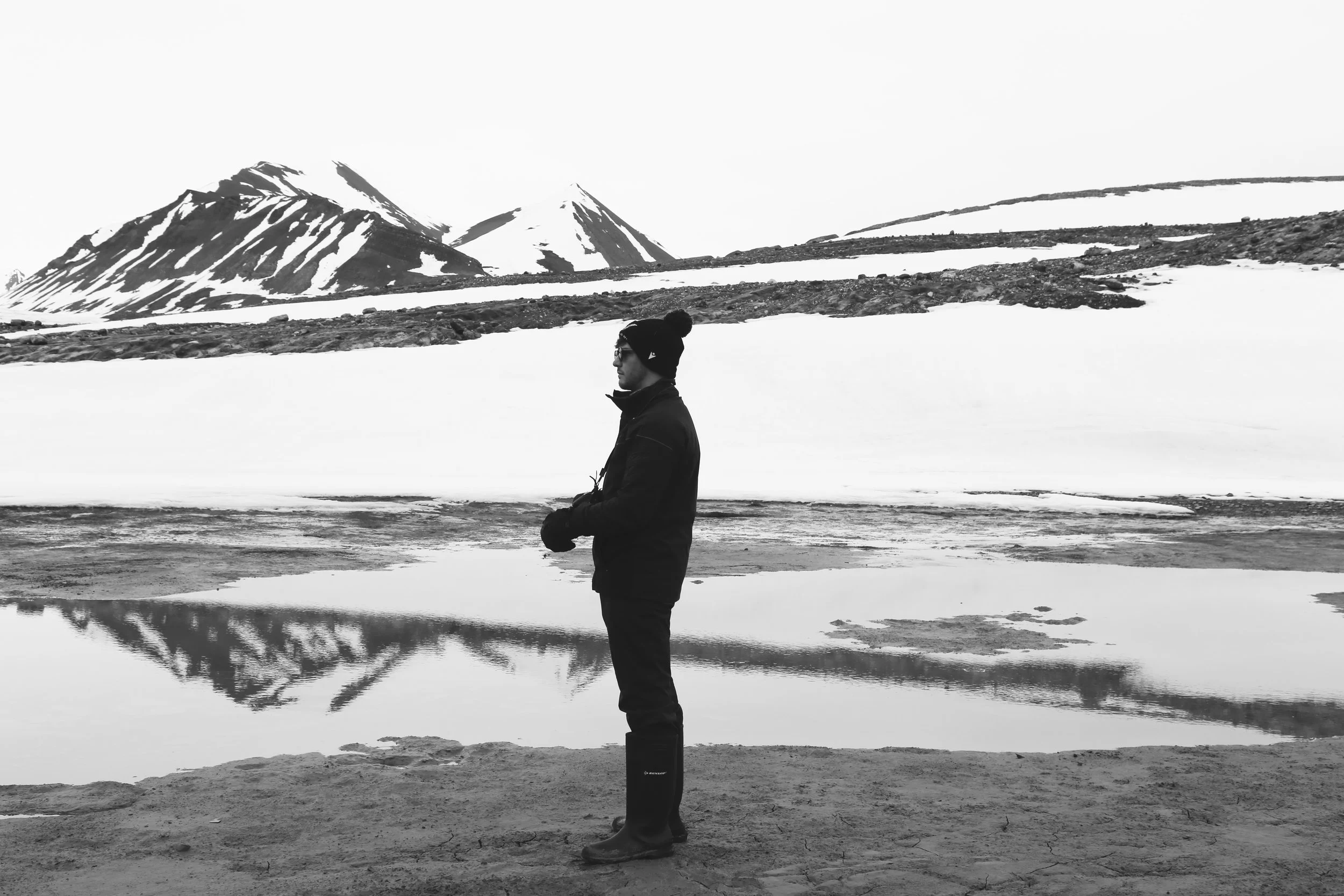A person dressed warmly in winter gear, including a beanie and rubber boots, standing on rocky ground near a partially frozen body of water with snowy mountains in the background.