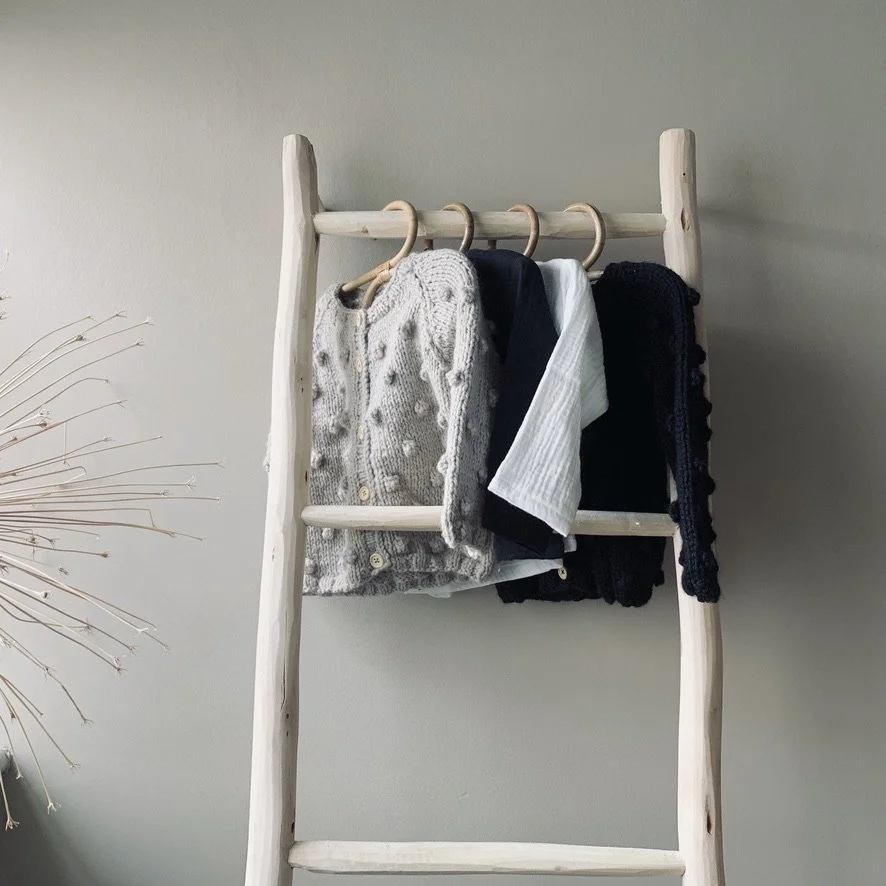 Clothes hanging on a wooden ladder against a plain wall, including a gray cardigan, a navy shirt, a white shirt, and a black scarf.