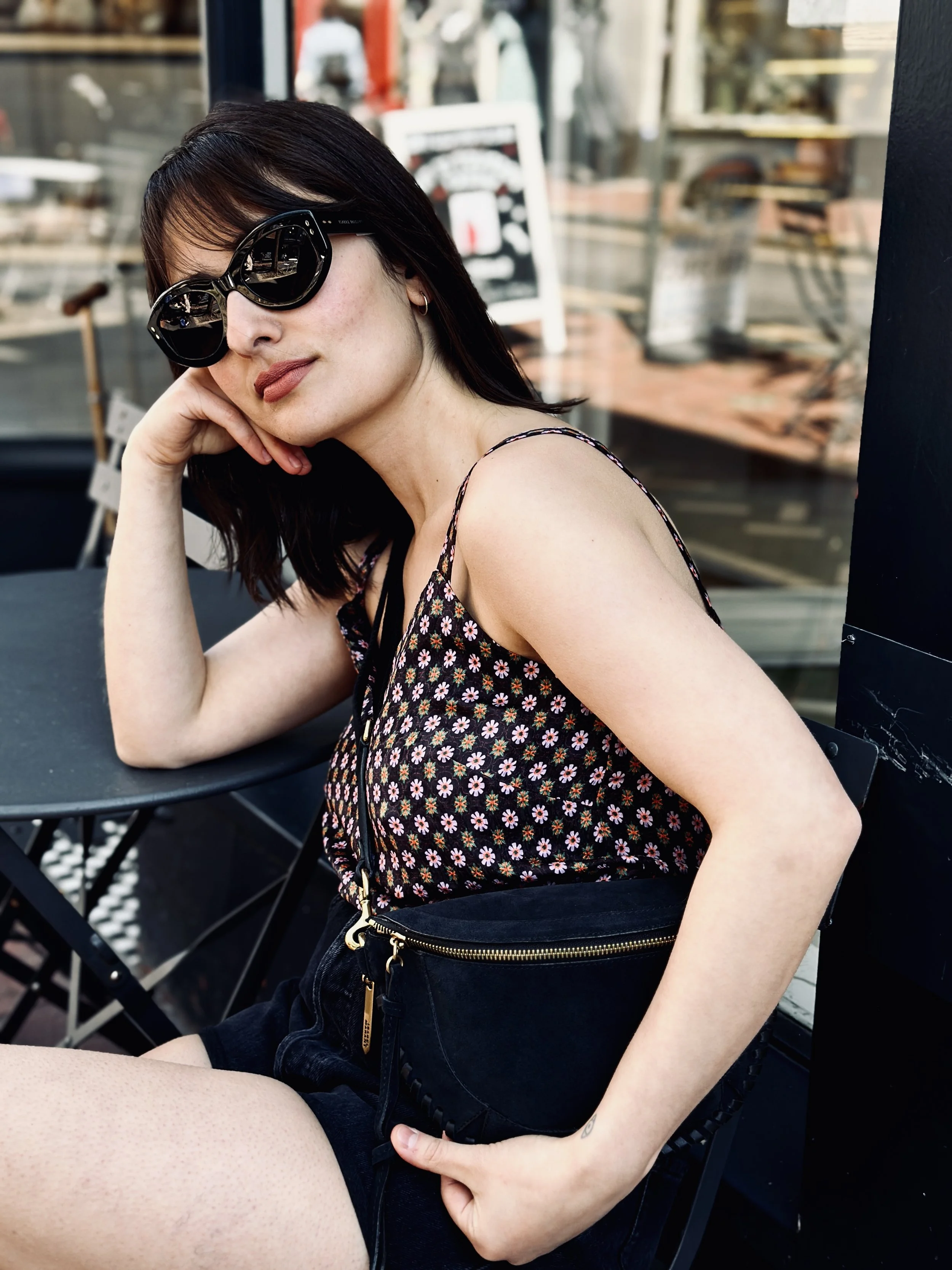 A woman with dark hair wearing large black sunglasses, a floral top, and black shorts sitting outside at a table with her head resting on her hand.