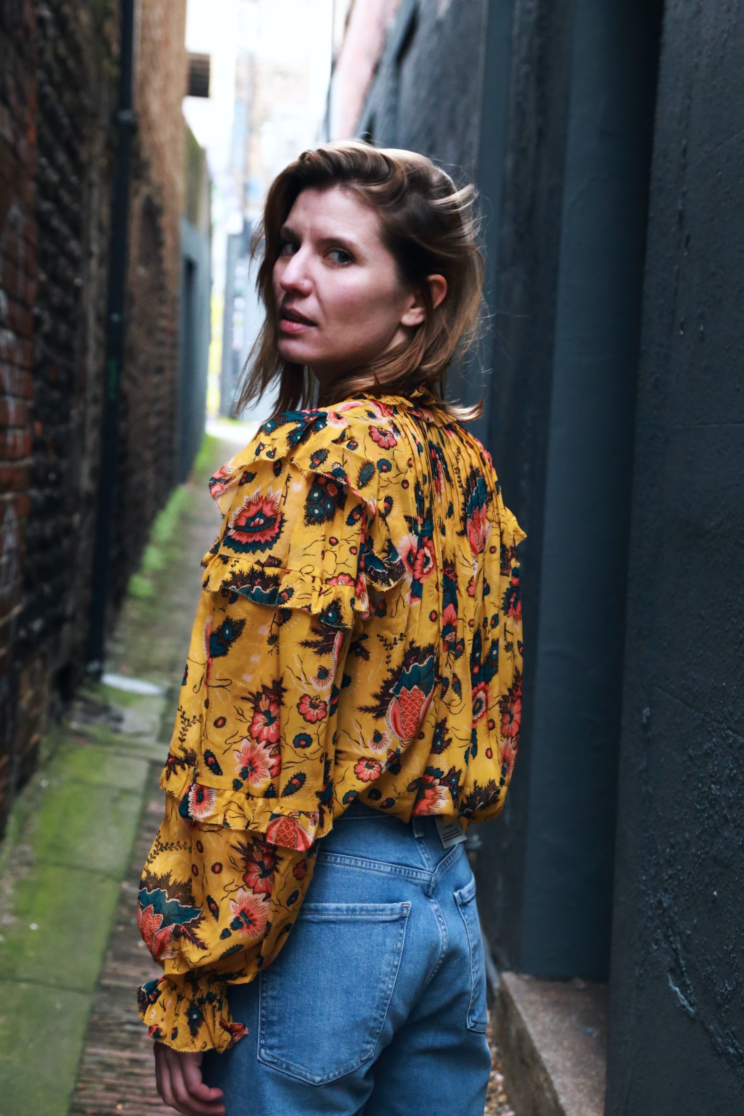 A woman with shoulder-length brown hair wearing a yellow floral blouse and blue jeans, standing in an alleyway with brick and painted walls.