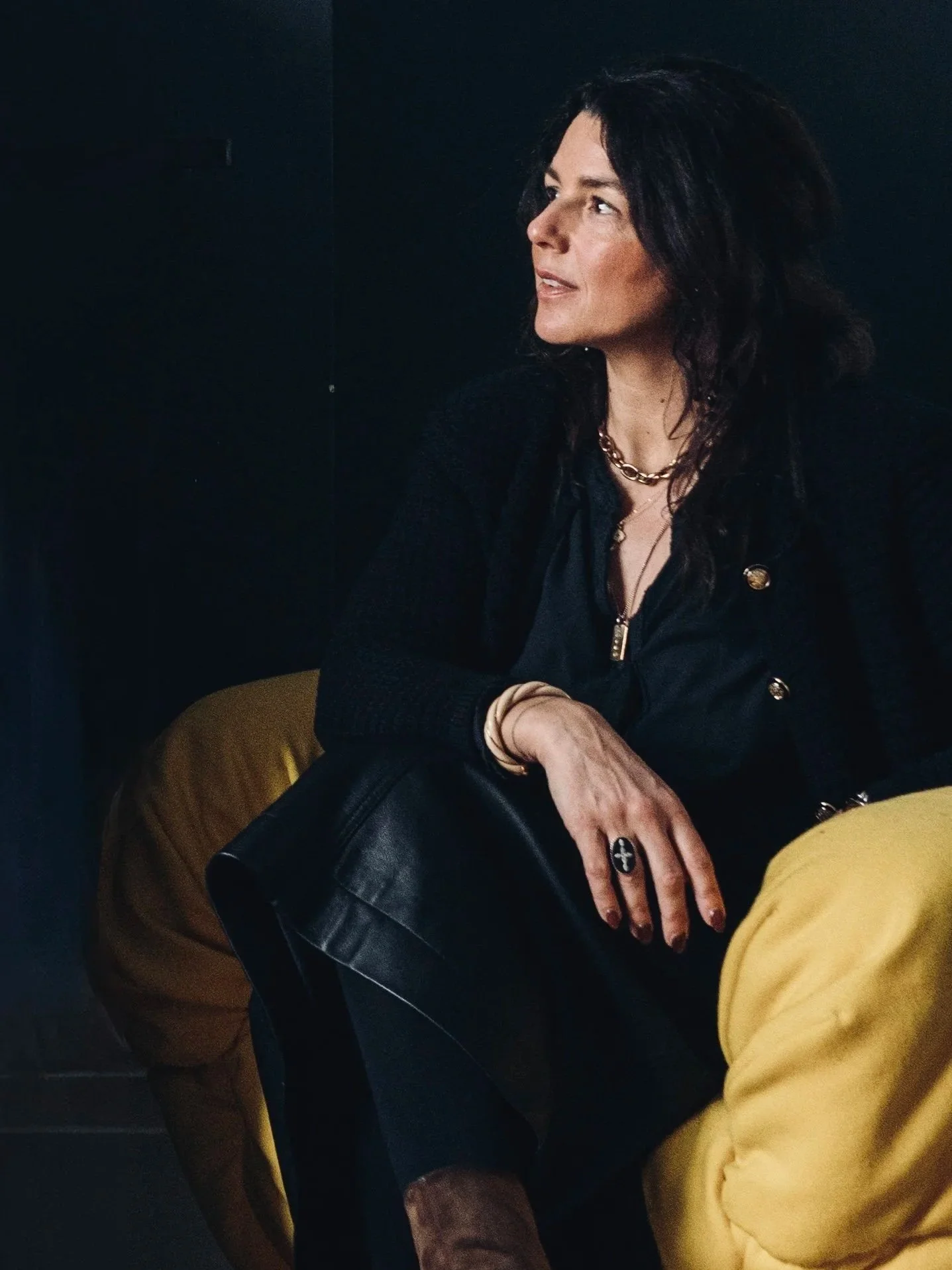 A woman sitting in a dark room, dressed in black with gold jewelry, holding a black purse, and wearing yellow pants