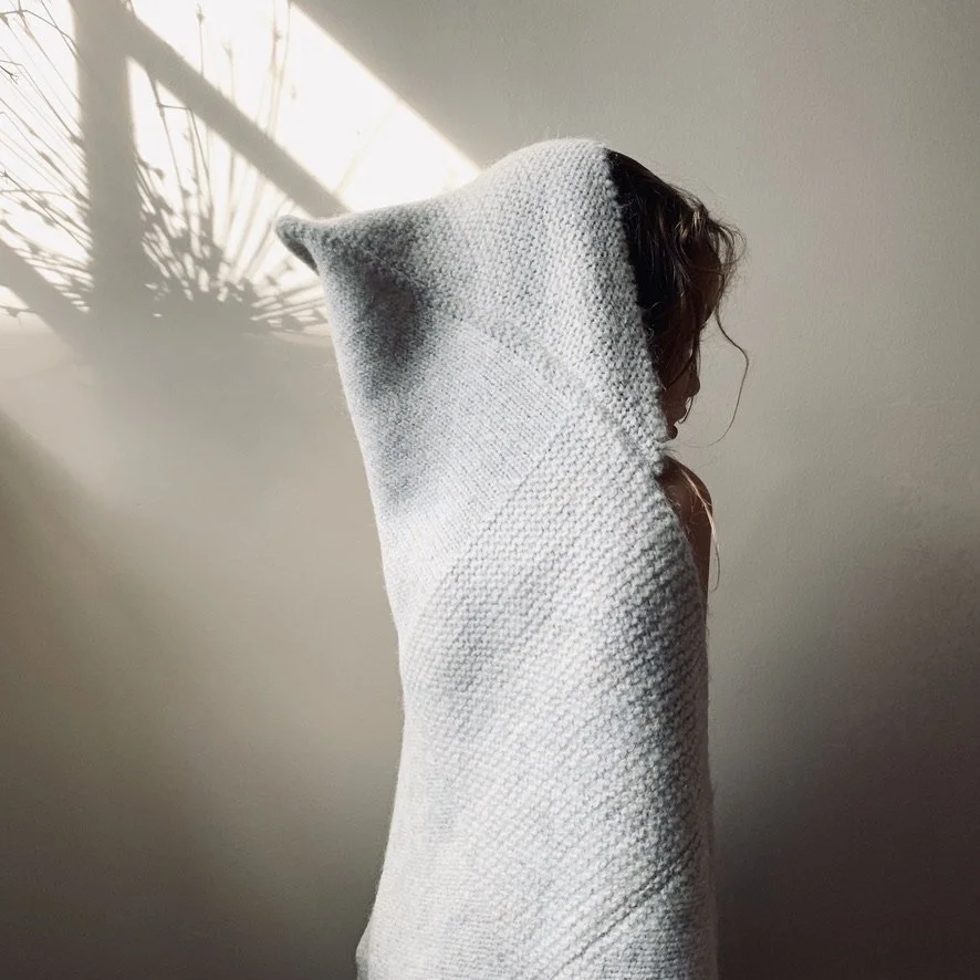 Person with long hair wearing a large, light gray hooded garment, standing against a plain wall with sunlight casting a shadow of plant leaves.