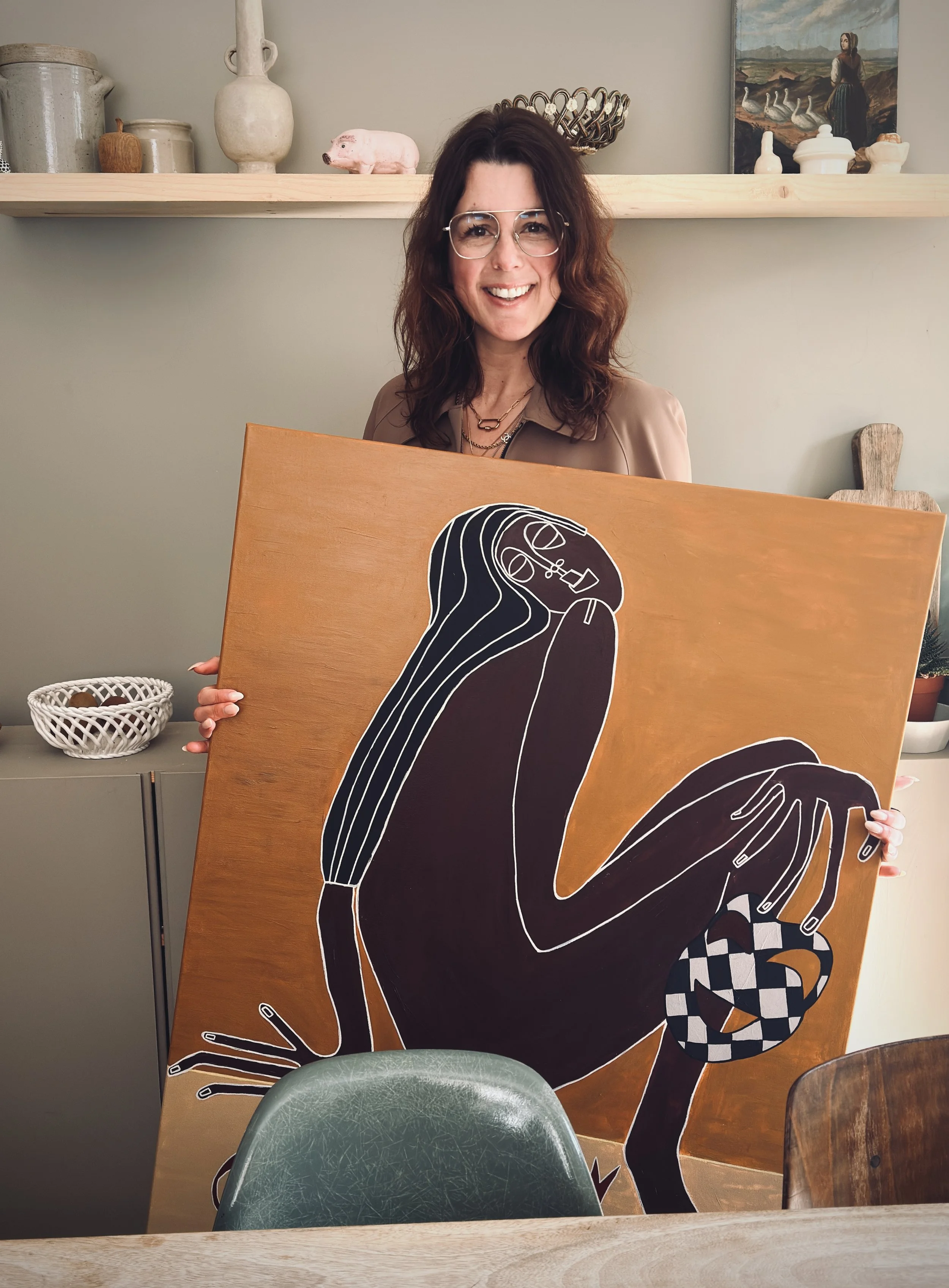 Woman with glasses smiling, holding a large artwork of a stylized woman with long hair, glasses, and long fingers, sitting on the ground, holding a checkered pumpkin.