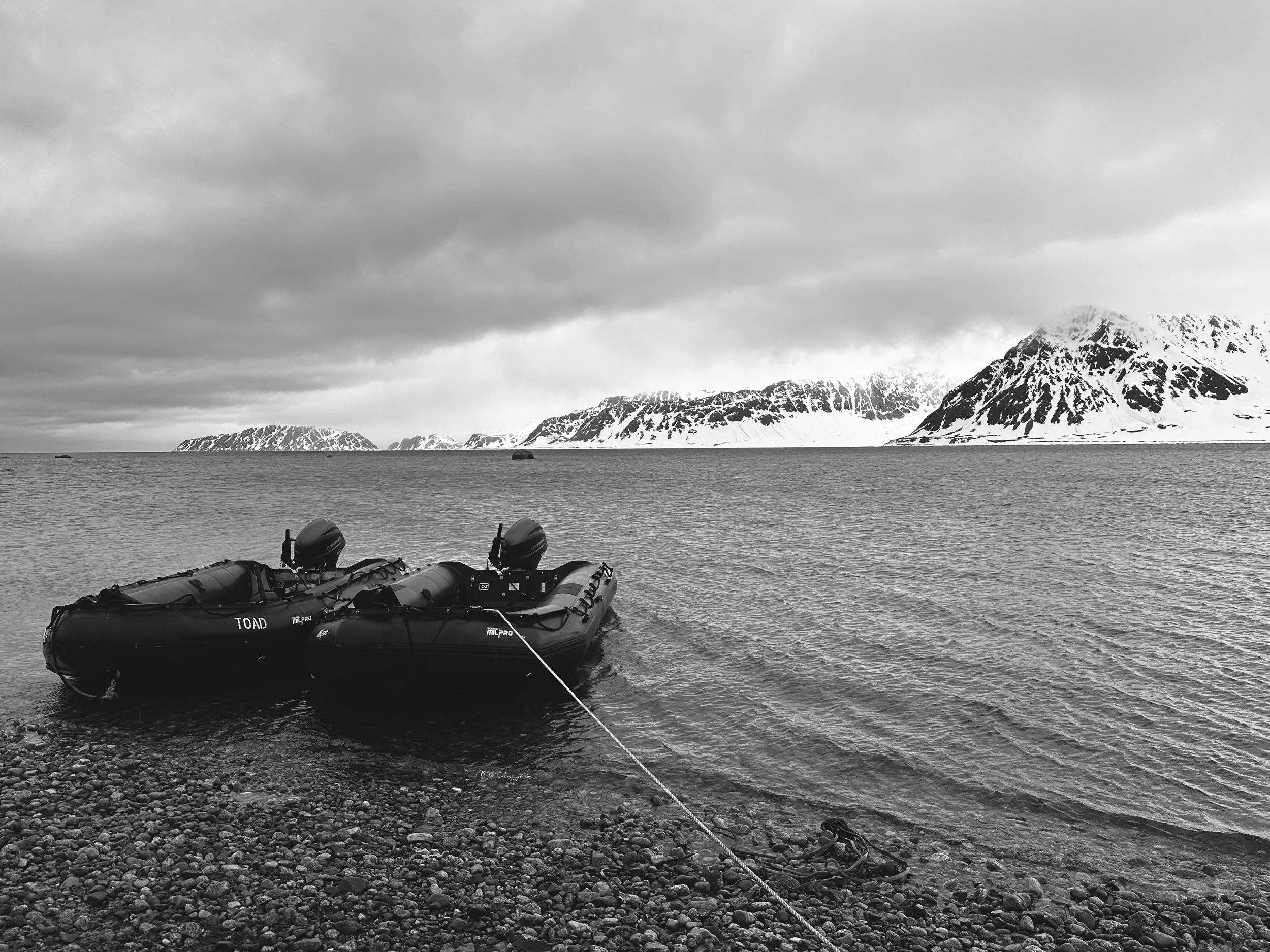 Inflatable boats tied to rocky shore with snow-covered mountains and cloudy sky in background.