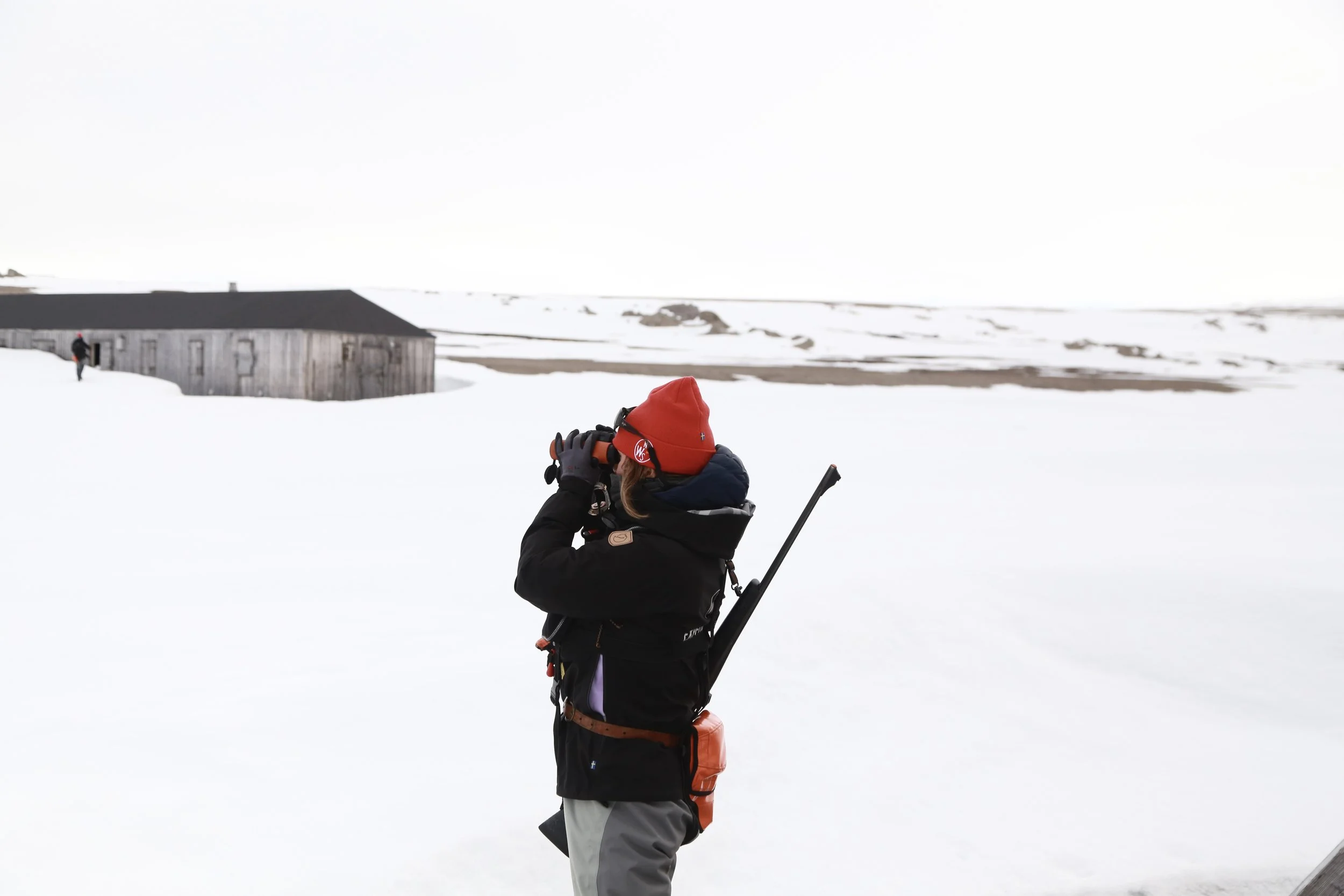 Person in winter gear with red hat using binoculars in snow-covered landscape with an old barn in the background.