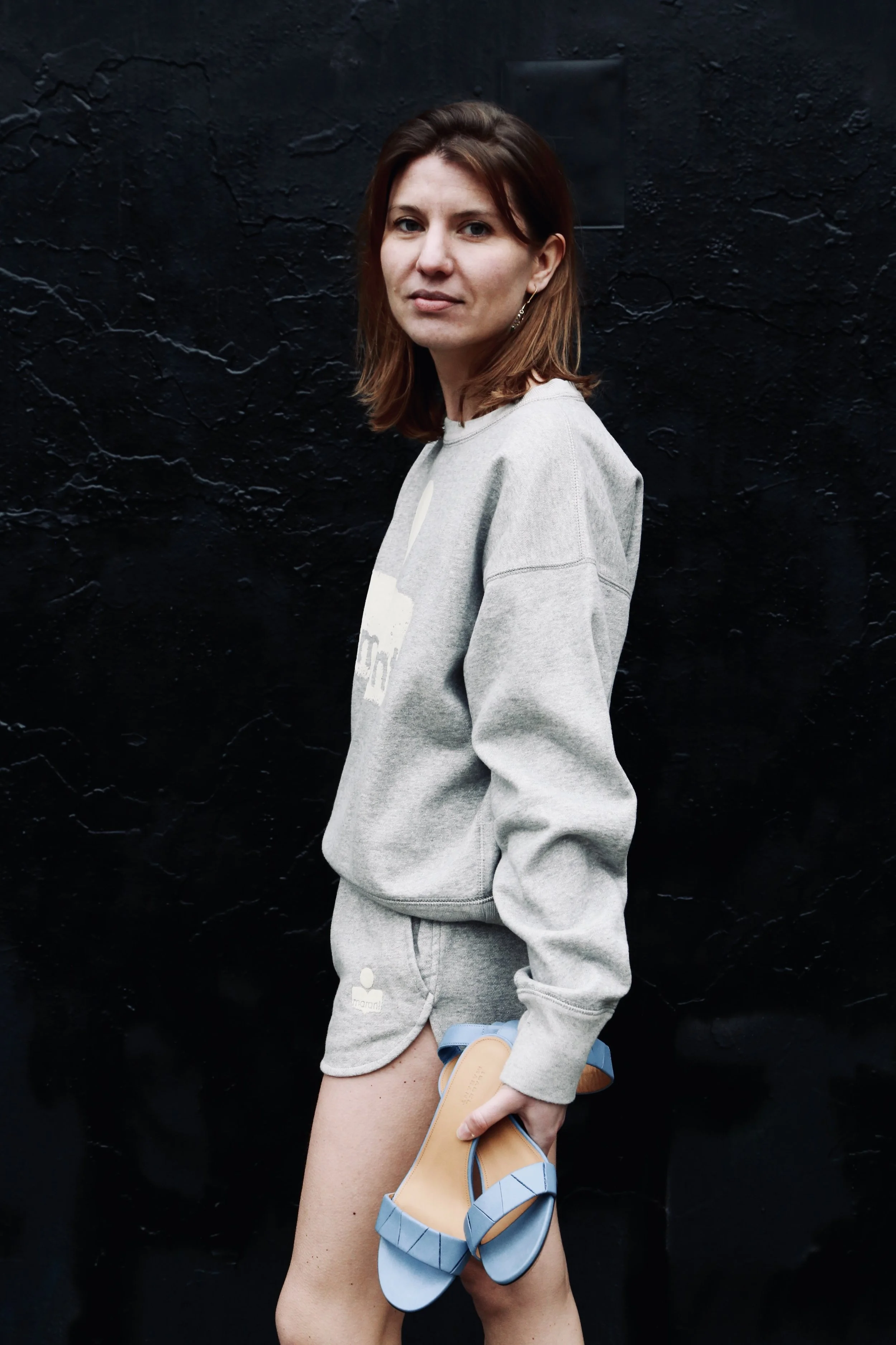 A young woman with brown hair, wearing a gray sweatshirt and matching shorts, is holding blue sandals against a black textured wall.