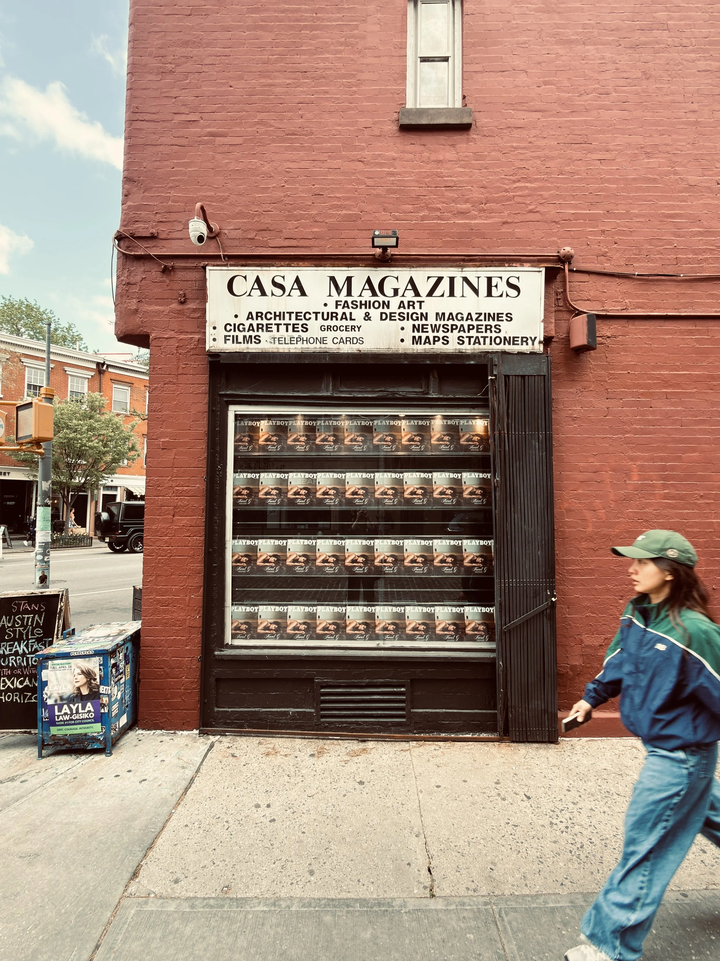 Casa Magazines newsstand on a red-brick corner, man in a green cap walking past, West Village