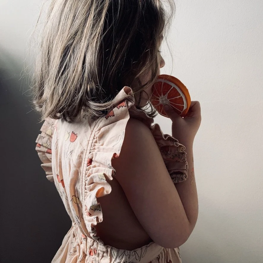 A young girl with shoulder-length hair wearing a ruffled, sleeveless dress with floral print, holding a slice of orange near her face, standing against a plain white wall.