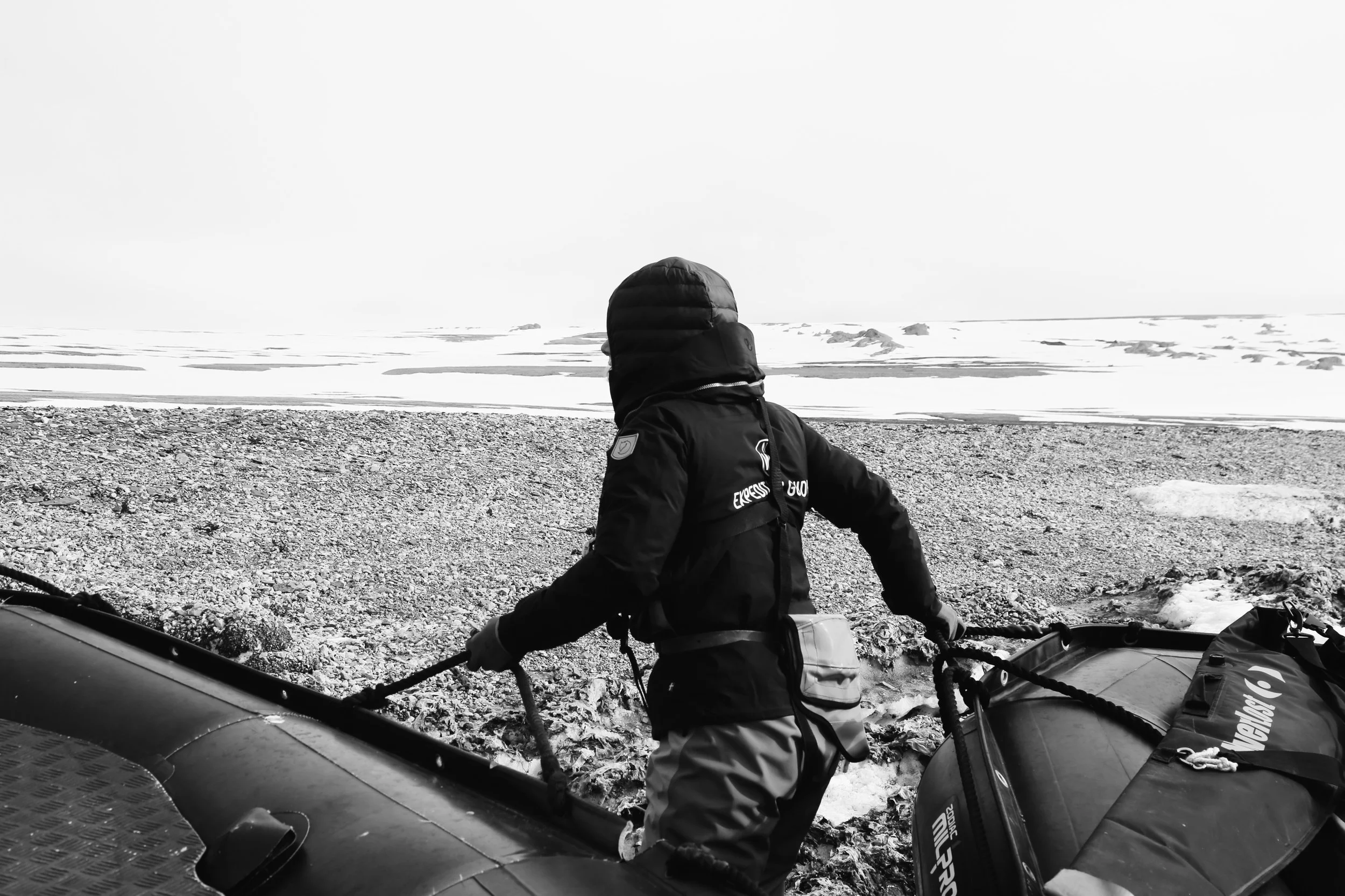 A person in winter gear, including a hooded jacket and gloves, prepares a rubber boat on a shore in a snowy, icy landscape.