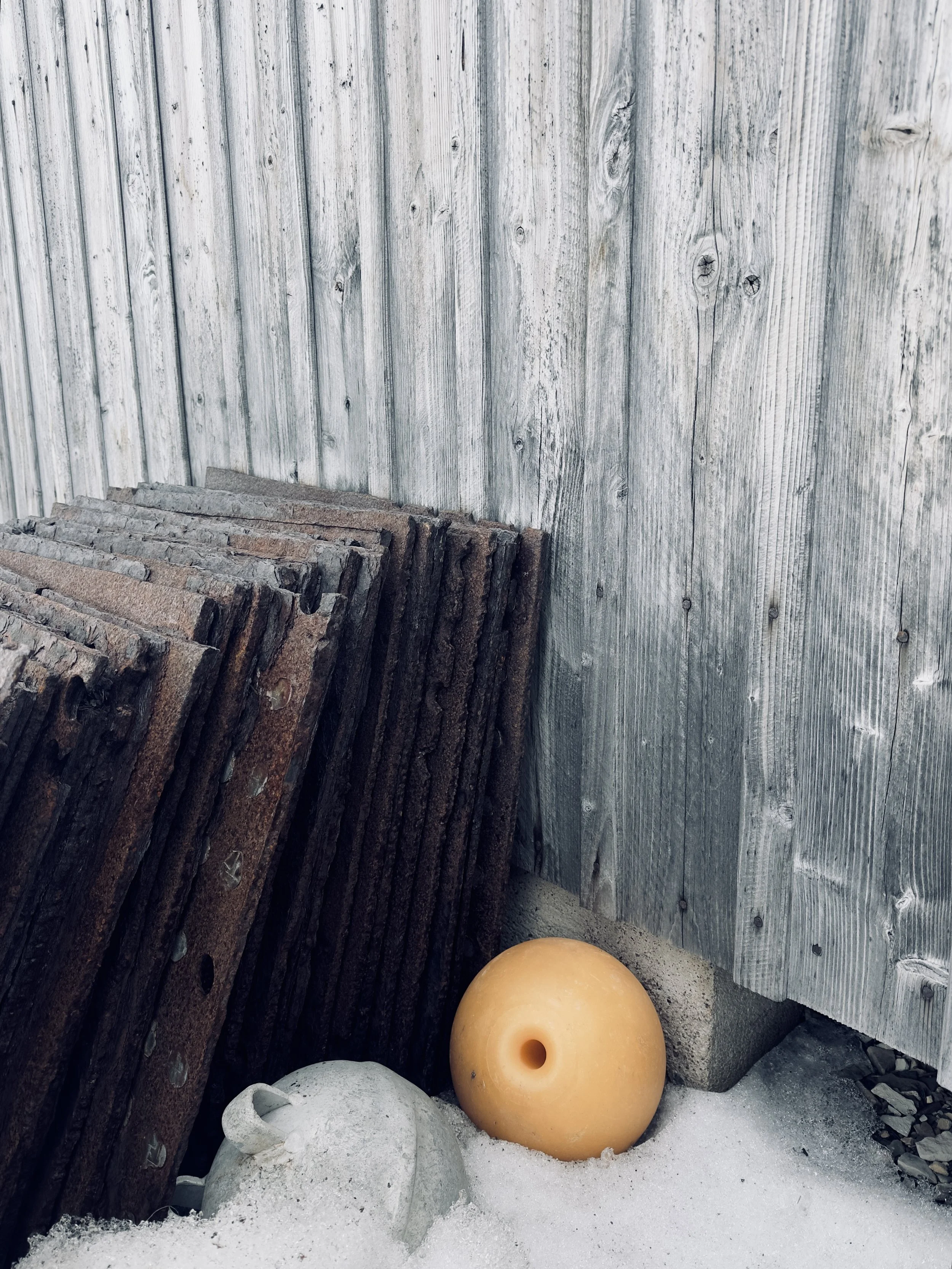 Stack of wooden railroad ties leaning against a weathered wooden fence, with a yellow buoy and a gray object on the sandy ground at the base of the fence.