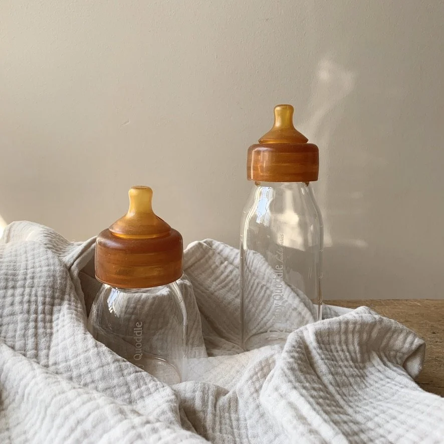 Two baby bottles with wooden caps and steam rising are placed on a white cloth on a wooden surface.