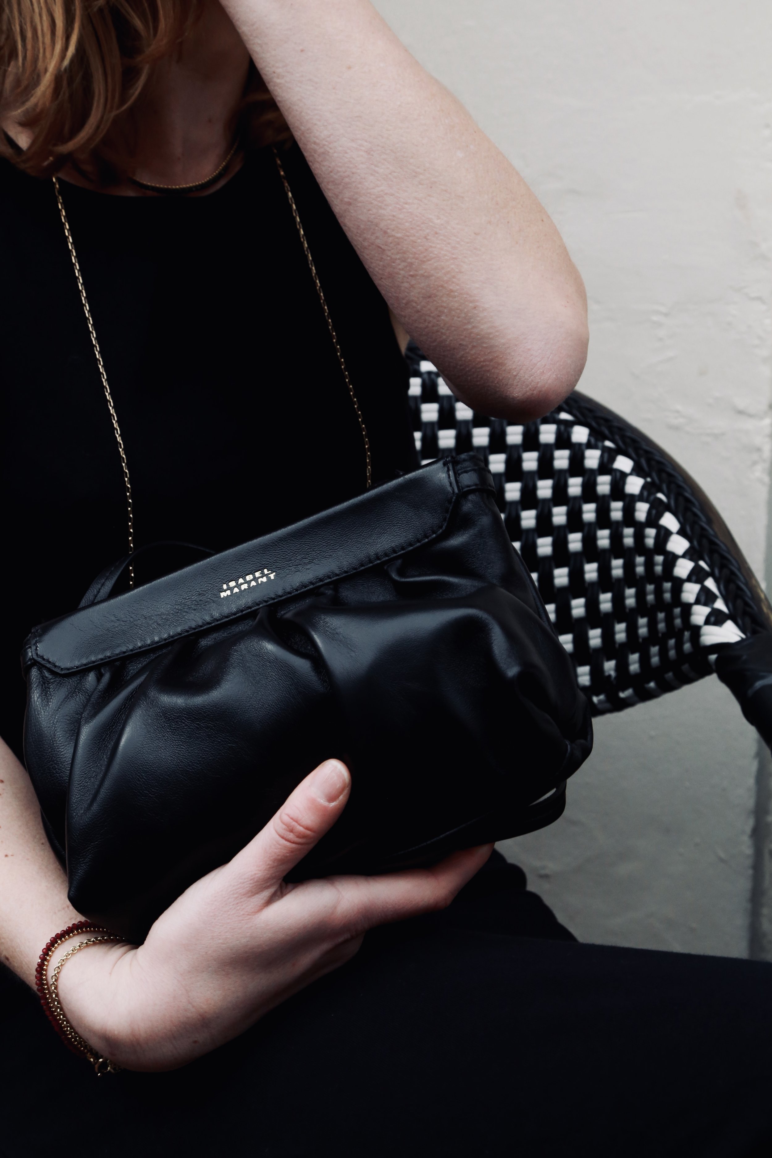 Person wearing a black top with a long gold chain necklace, holding a black leather Marc Marant purse, sitting on a chair with a black and white woven pattern backrest, against a light-colored wall.