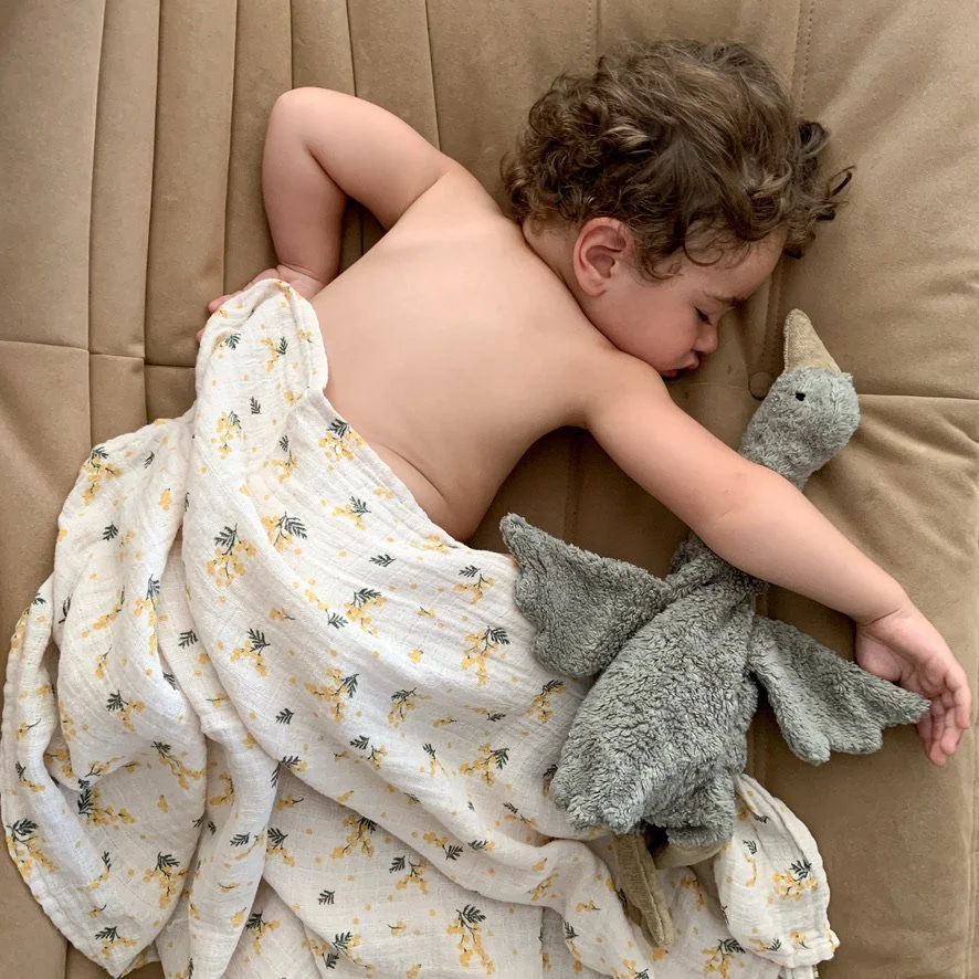 A young child with curly hair sleeping on a beige couch, wrapped in a white blanket with yellow and black floral patterns, holding a grey stuffed elephant toy.