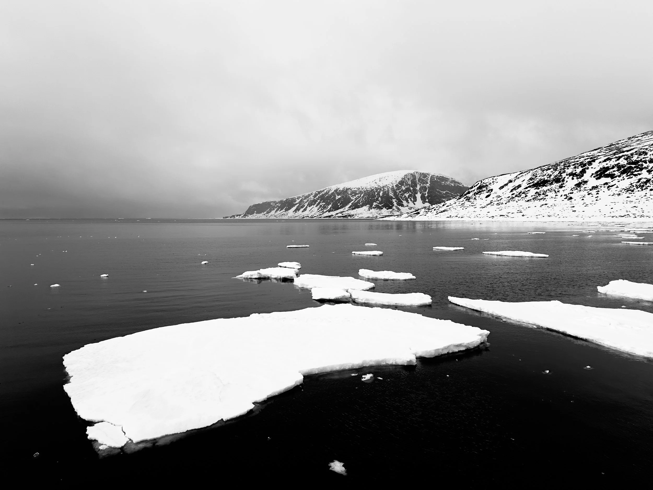 Black and white photo of ice floating on a calm body of water with snowy mountains in the background and cloudy sky overhead.