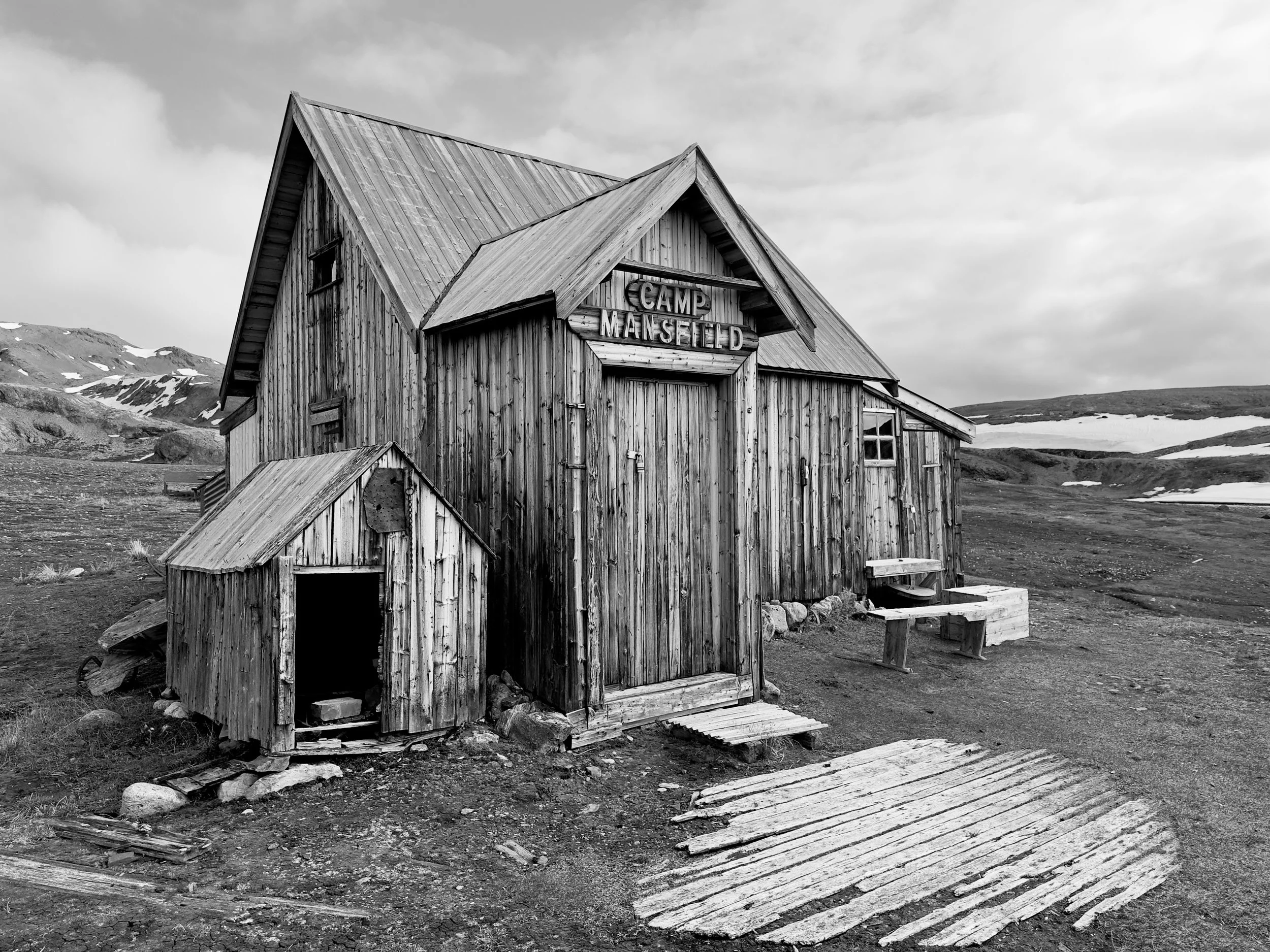 A weathered wooden cabin labeled 'Camp Mansfield' in a barren landscape with snow patches and mountainous terrain in the background.