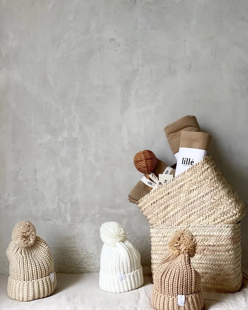 Three knitted hats with pom-poms in beige, white, and tan, placed on a white surface with a woven basket filled with rolled-up fabric or textiles and a small decorative wicker item, against a light gray textured wall.