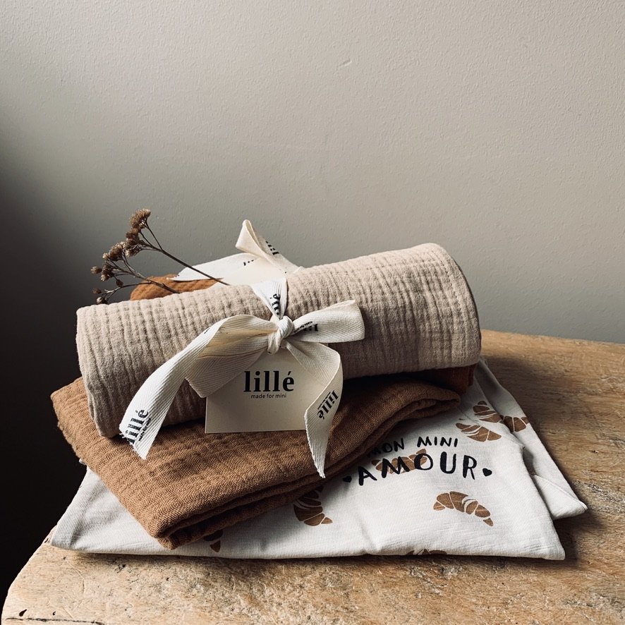 A neatly folded beige and brown fabrics or towels with a white ribbon and a tag that reads "lillé made for mini" resting on a wooden surface, next to a white cloth with brown text and leaf patterns.