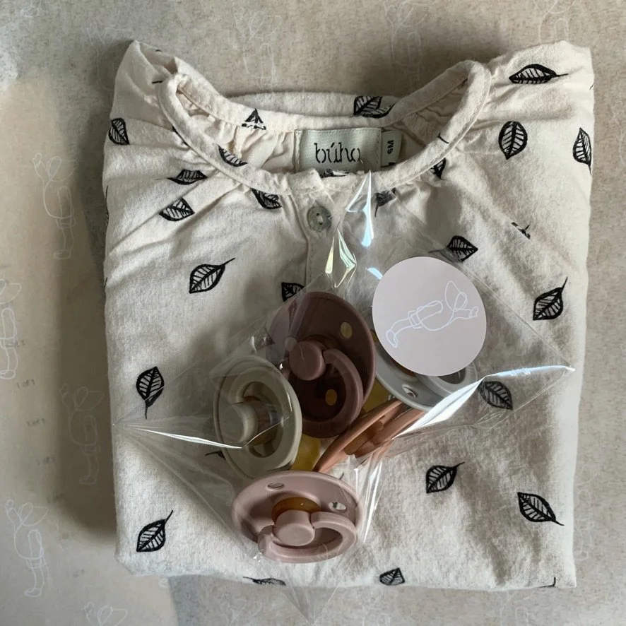 A folded shirt embroidered with black leaves, with a tag reading 'bubu', partially covered by a plastic bag containing pastel-colored pacifiers.