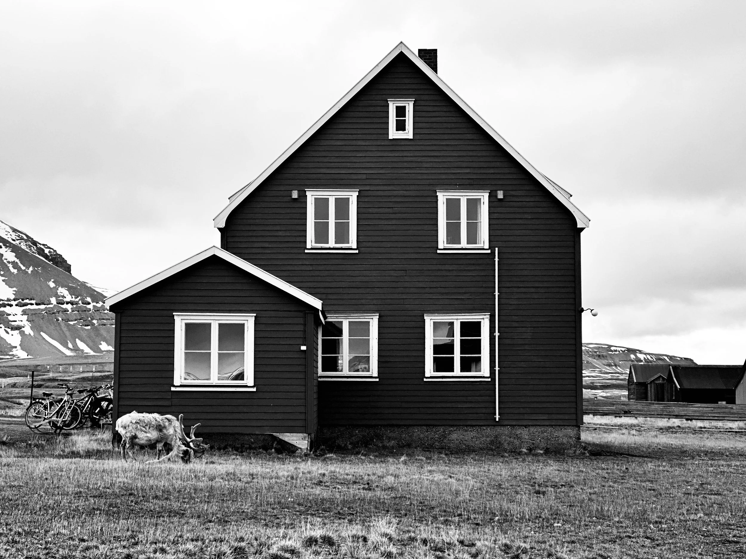 A black house with white window frames and a small attached shed, situated in a rural area with mountains in the background. There are bicycles and a reindeer grazing in the foreground.